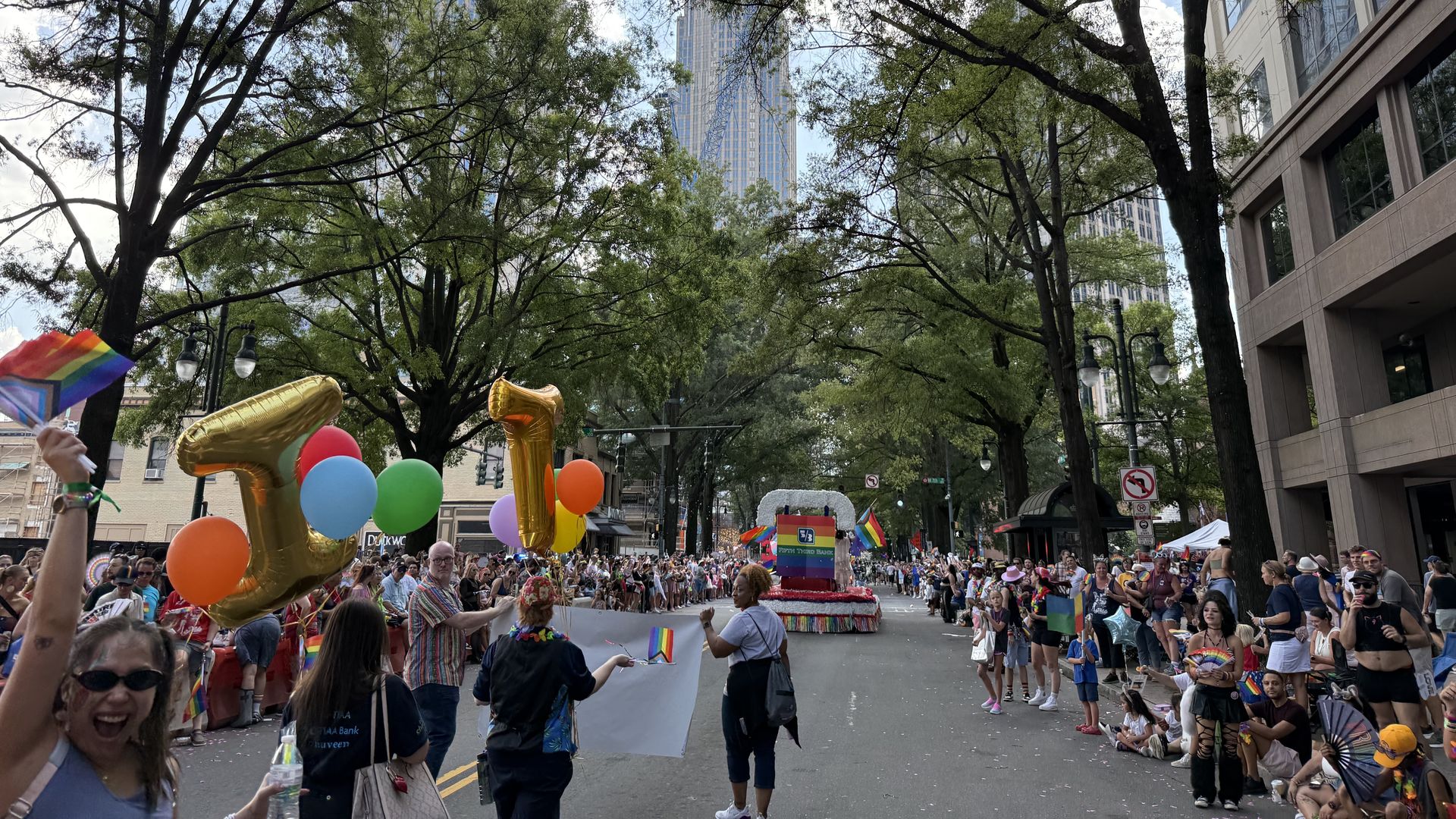 Busy city street parade scene with a large crowd, rainbow flags, and colorful balloons. Participants march along a tree-lined avenue as tall buildings loom in the background.