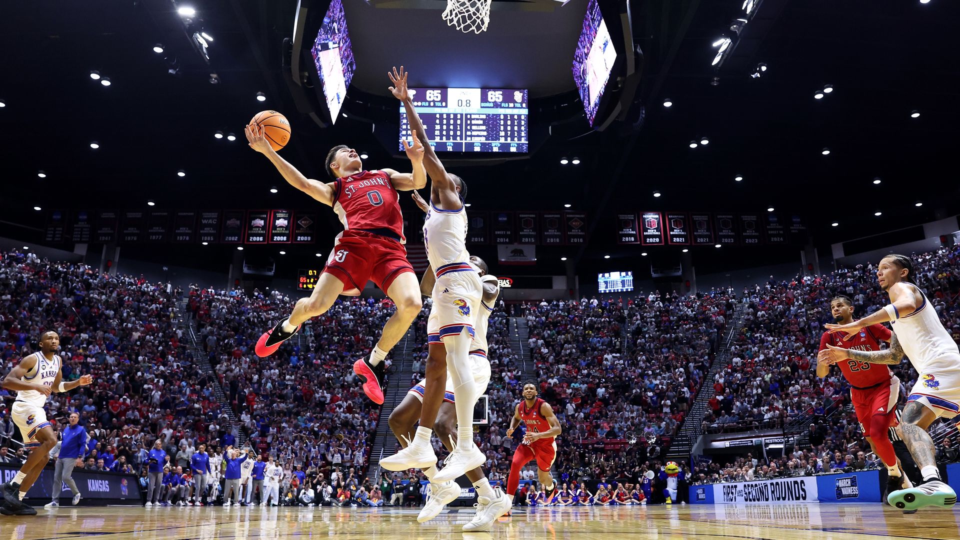 A view from the floor of Dylan Darling #0 of St. John's Red Storm players going up for a layup against a Kansas Jayhawks player during a 2026 NCAA Men's Basketball Tournament game with fans packed in the stands at Viejas Arena. 