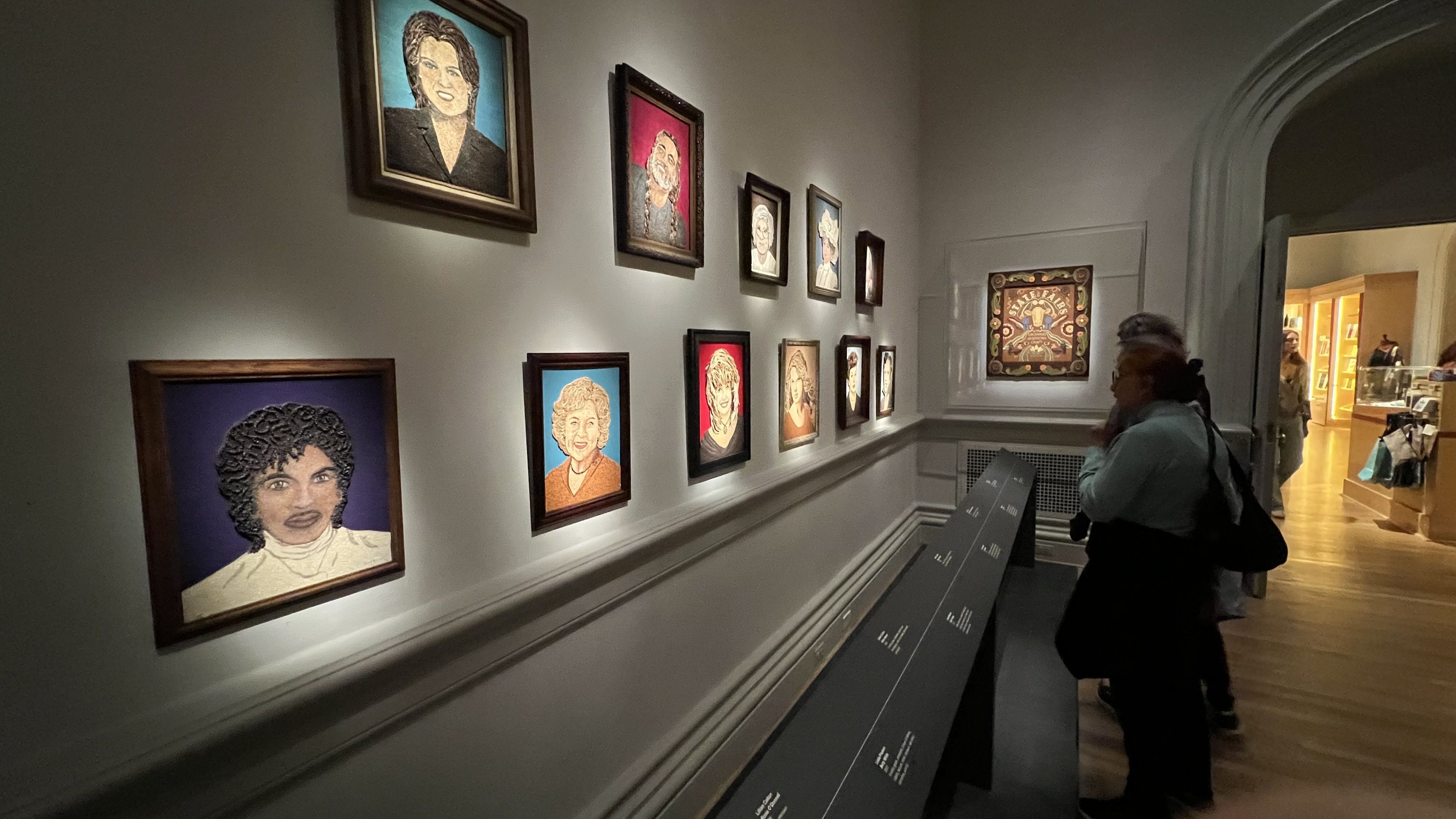 Gallery room with lighted portraits framed on the wall, showing various faces with colorful backgrounds, two people closely viewing the artwork beside a display panel.