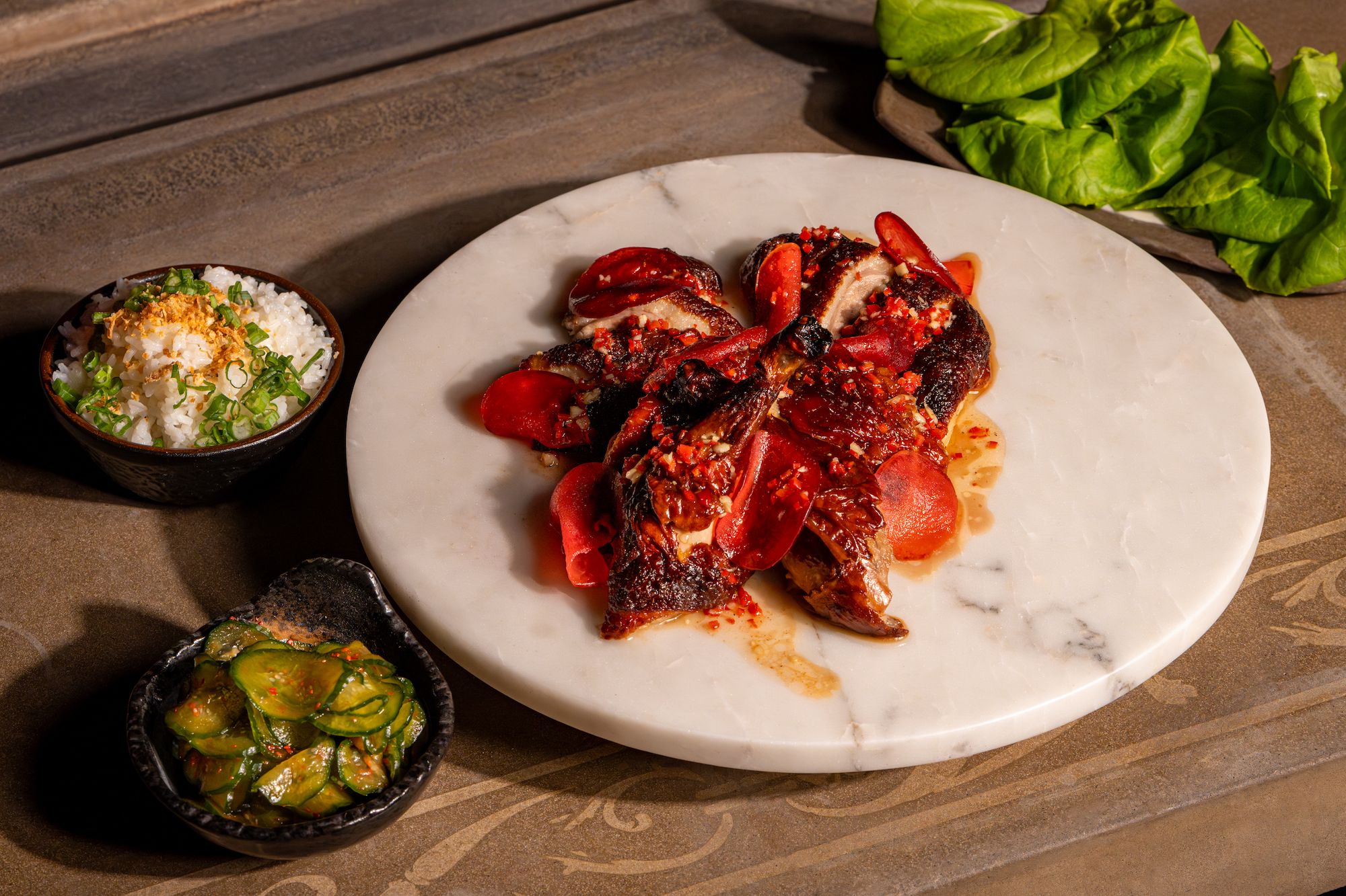 A marble platter of duck with side dishes of cucumbers and rice