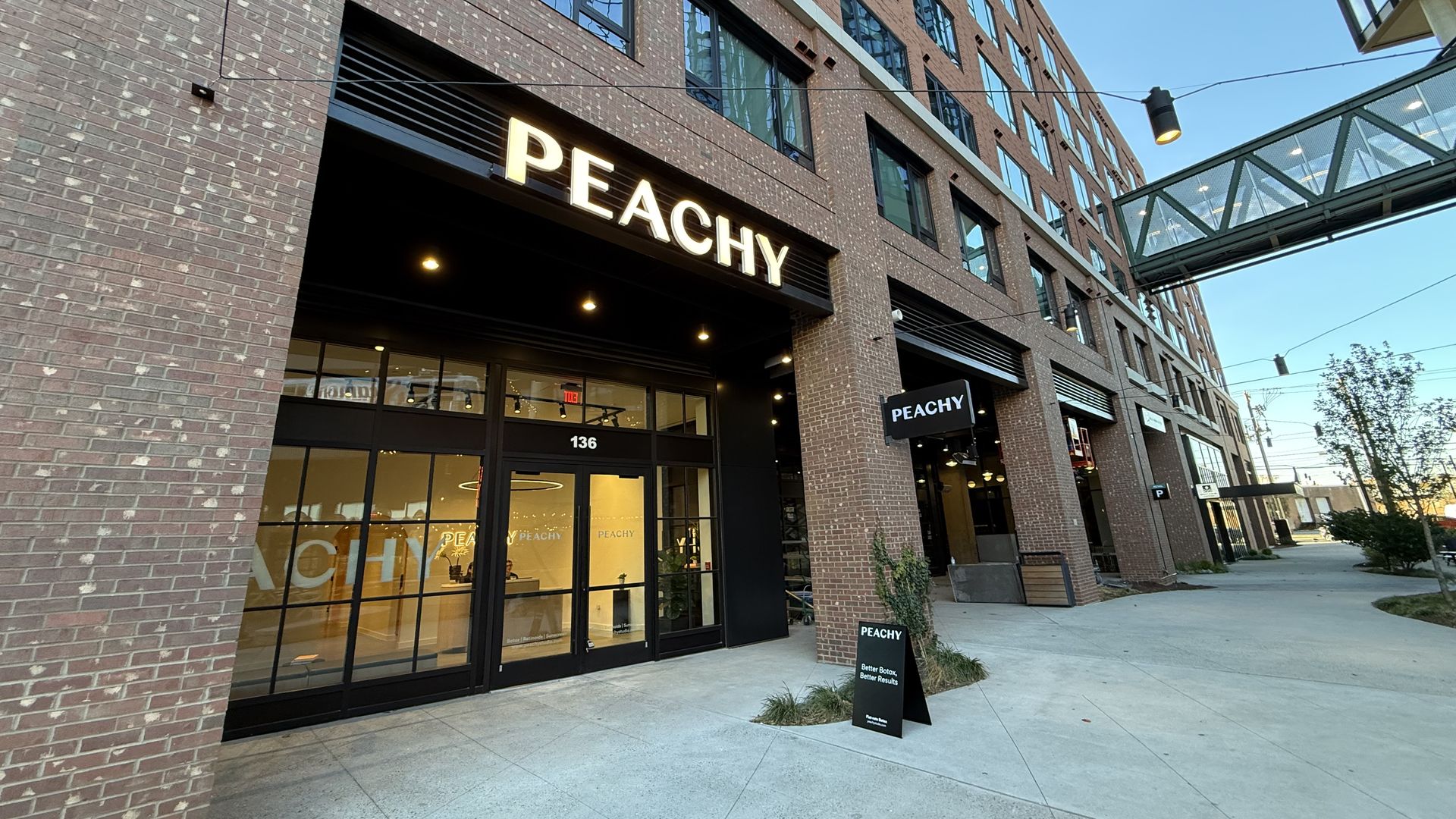 Brick storefront with large glass windows and a bright illuminated "PEACHY" sign. Modern multi-story brick building with an elevated glass skybridge and blue sky above.
