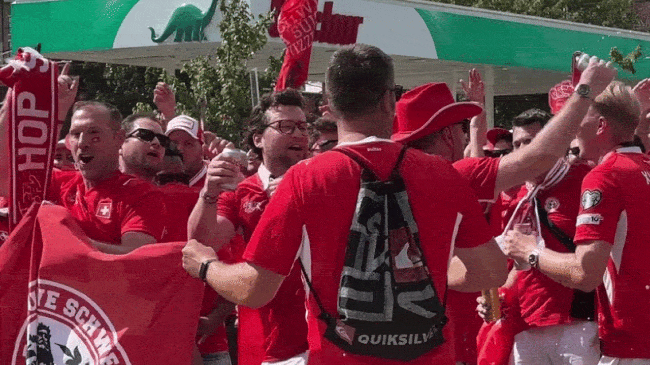 Group of red-clad fans cheering outdoors, holding drinks and flags; central figure with back to camera wears a black Quiksilver backpack amid the celebration.