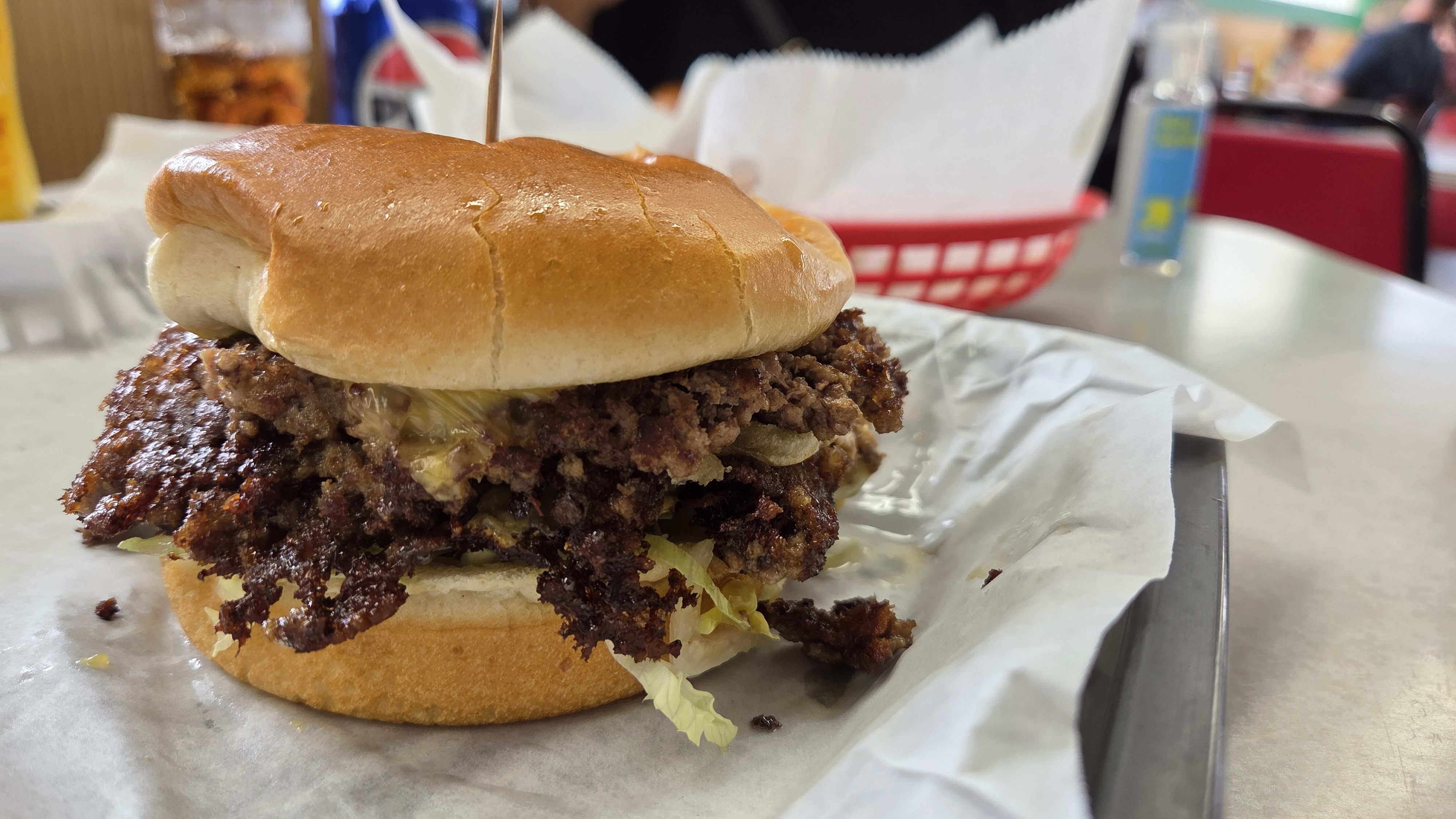 Close-up of a large crispy fried chicken sandwich on a glossy bun with lettuce, served on parchment in a casual diner; a red basket and a blue drink bottle blur in the background.