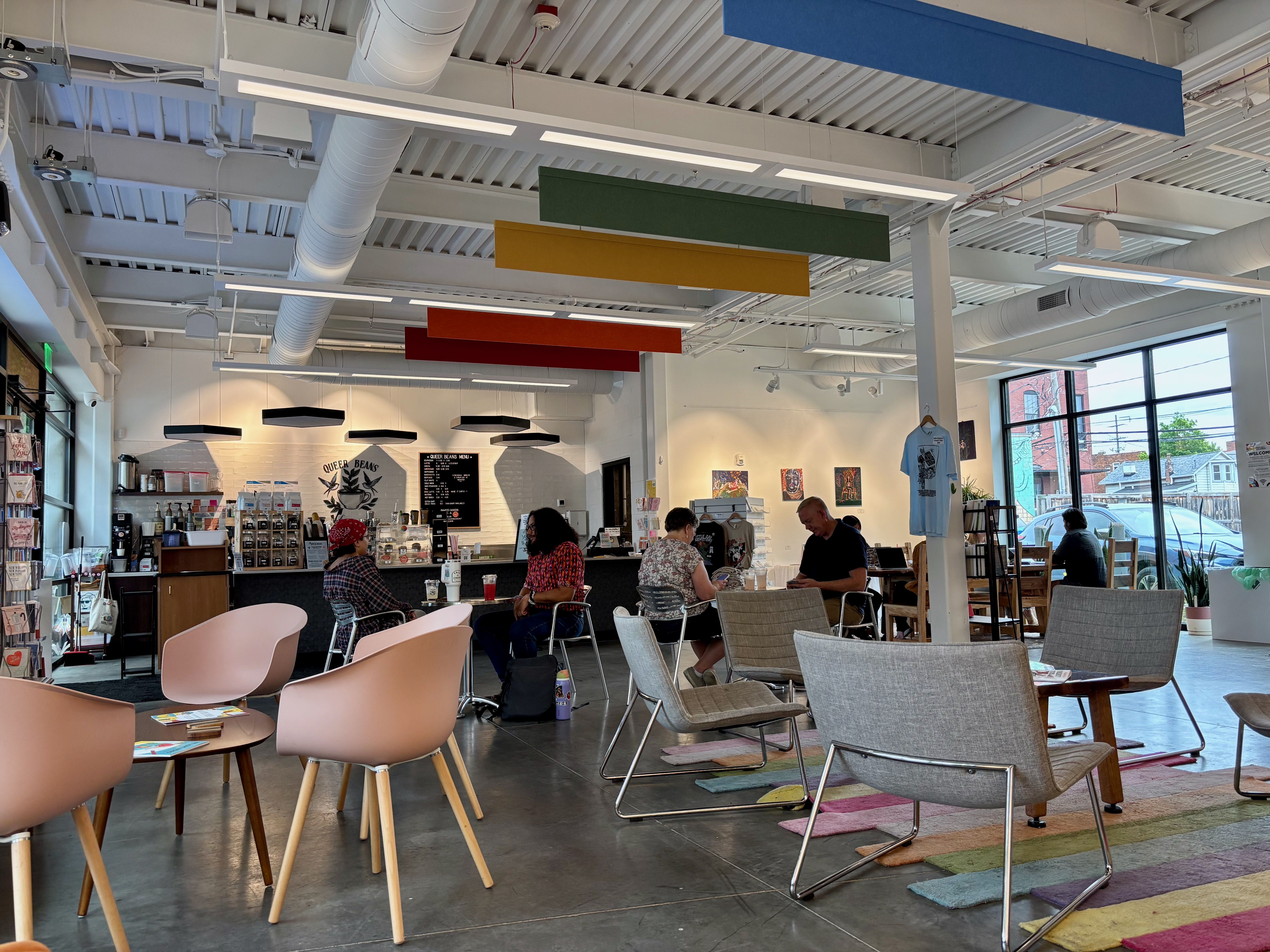 A bright, modern cafe with a white industrial ceiling and exposed ducts. Colorful rectangular hanging panels in blue, green, yellow, orange, and red float above tables where customers sit by large windows.
