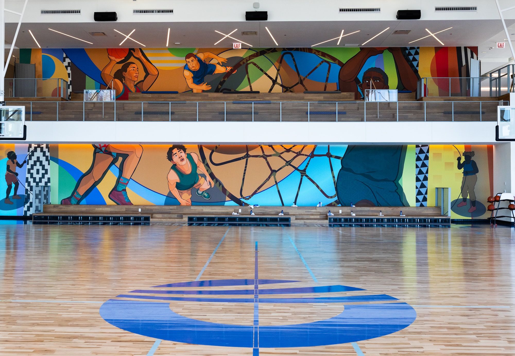 Indoor gym with a colorful mural of athletes along the back wall, two levels of seating, and a polished wooden basketball court featuring a large blue circle logo at center.