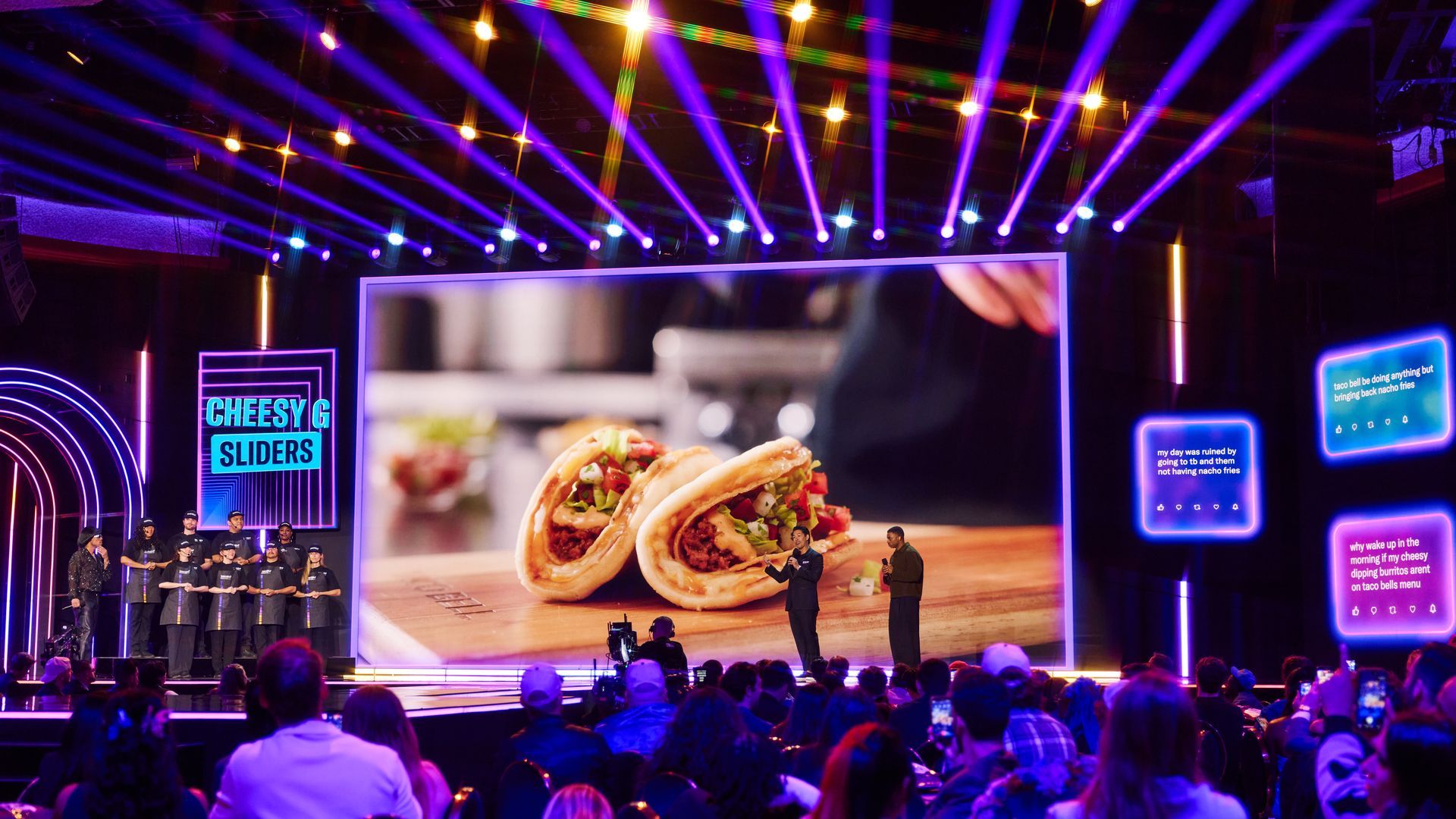 Vibrant stage with purple/blue laser lights; a large screen shows two loaded tacos on a wooden board. Two presenters speak at center as a group of chefs stands to the left; audience watches.