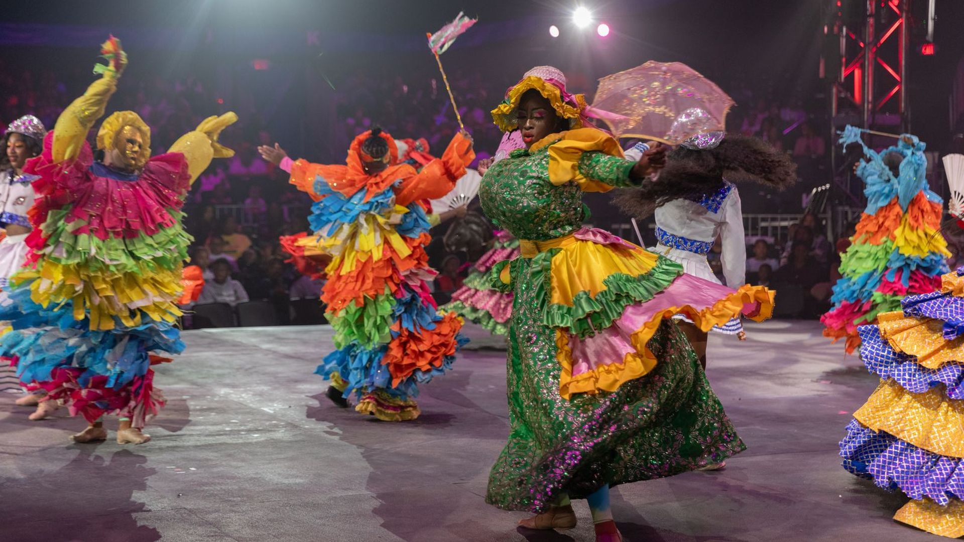 Vibrant stage scene: dancers in rainbow, multi-layered, ruffled dresses swirl and spin; bright spotlights, fans, and a seated audience in the dim background.