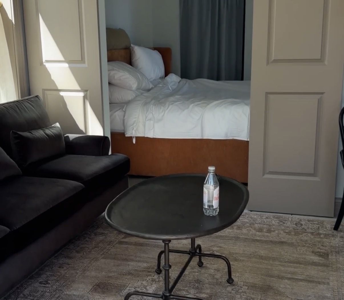 Living room with a black sofa on the left, a round coffee table holding a water bottle, and a neatly made white bed with an orange headboard in a small alcove, framed by beige double doors.