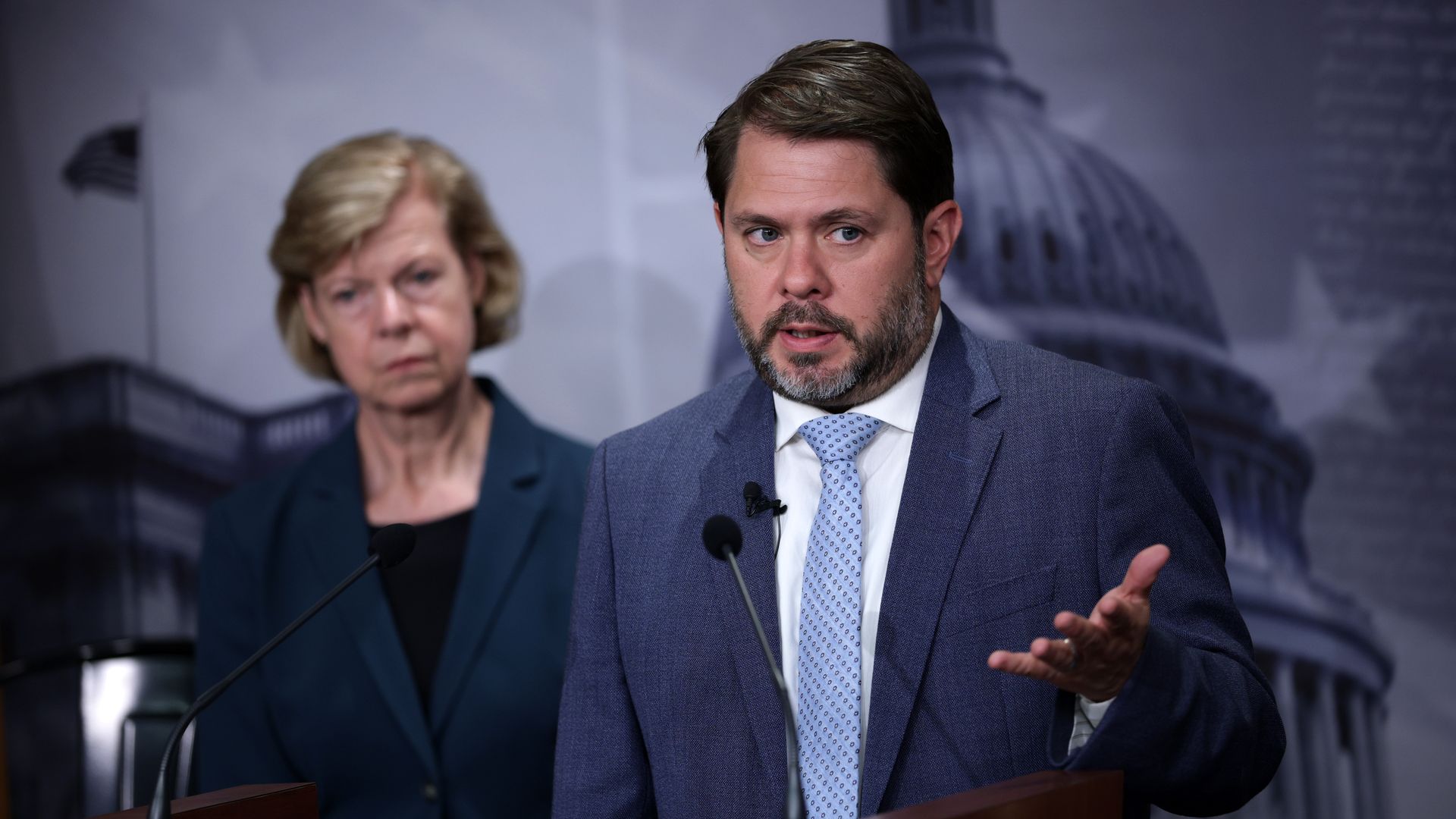 Man in a blue suit and light patterned tie speaks at a wooden podium with microphones, gesturing as an older woman in a dark blazer looks on in the background.