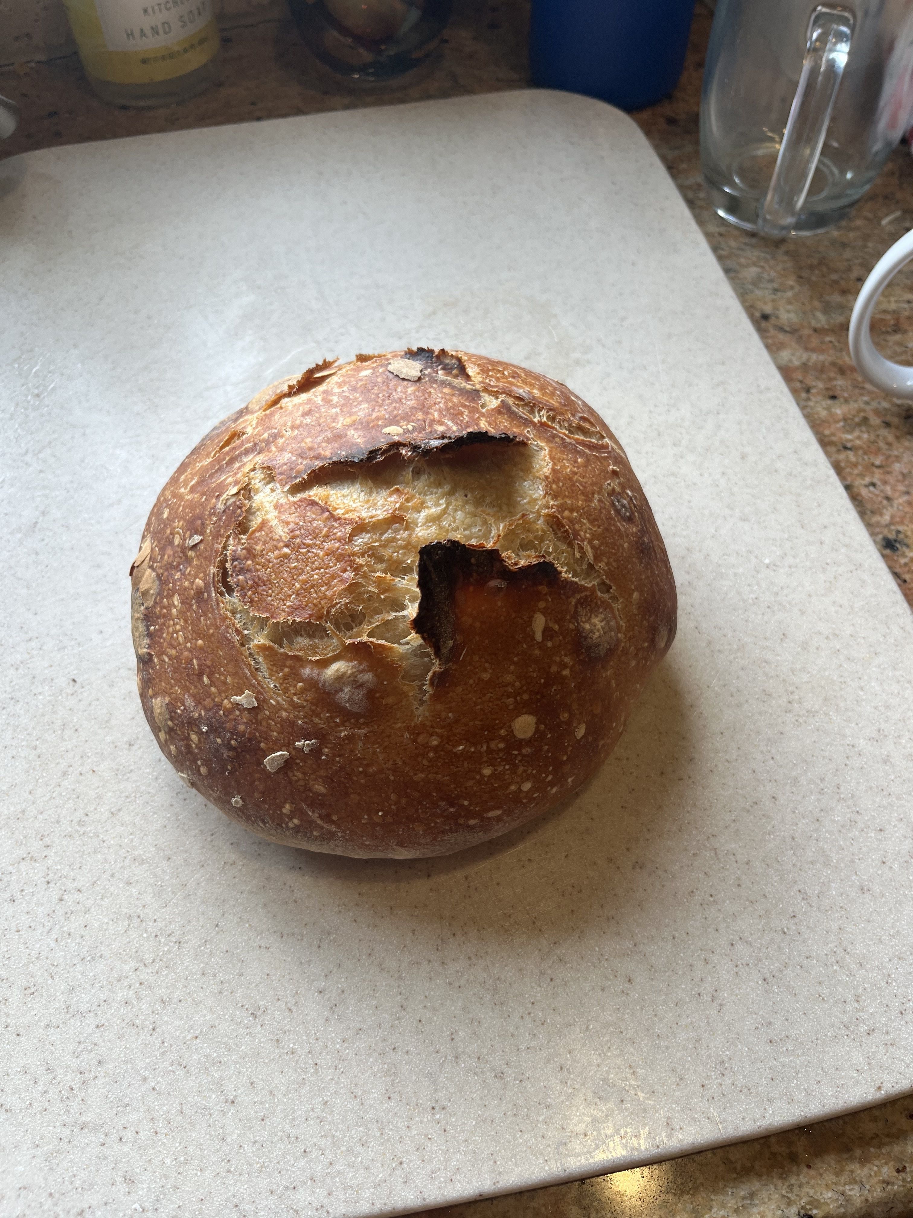 Round loaf of golden-brown crusty bread with cracks on top, placed on a beige speckled cutting board on a kitchen counter.