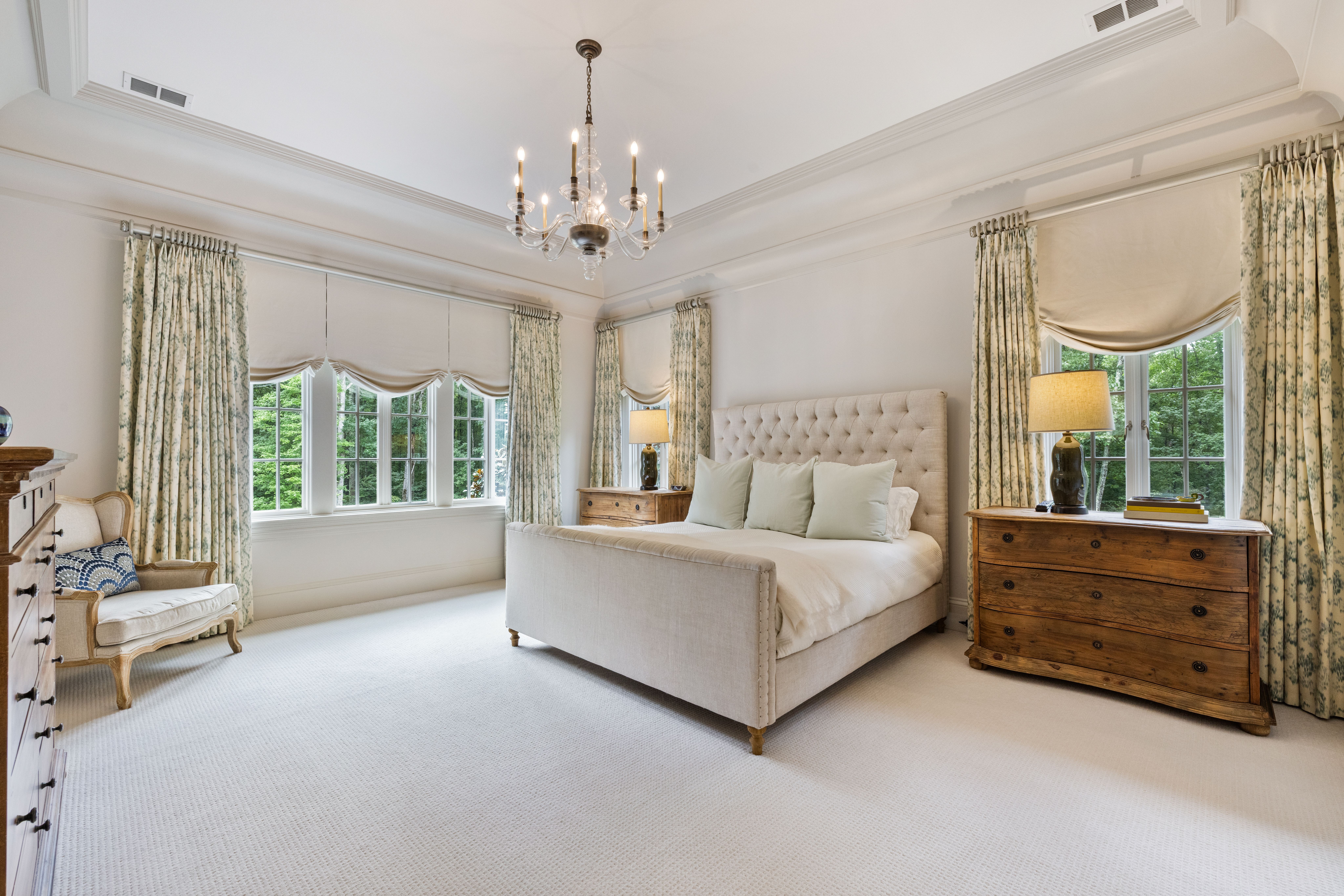 Bright bedroom with beige tufted bed, white bedding, wooden dressers with lamps, floral curtains, large windows, cream carpet, and a chandelier ceiling light.