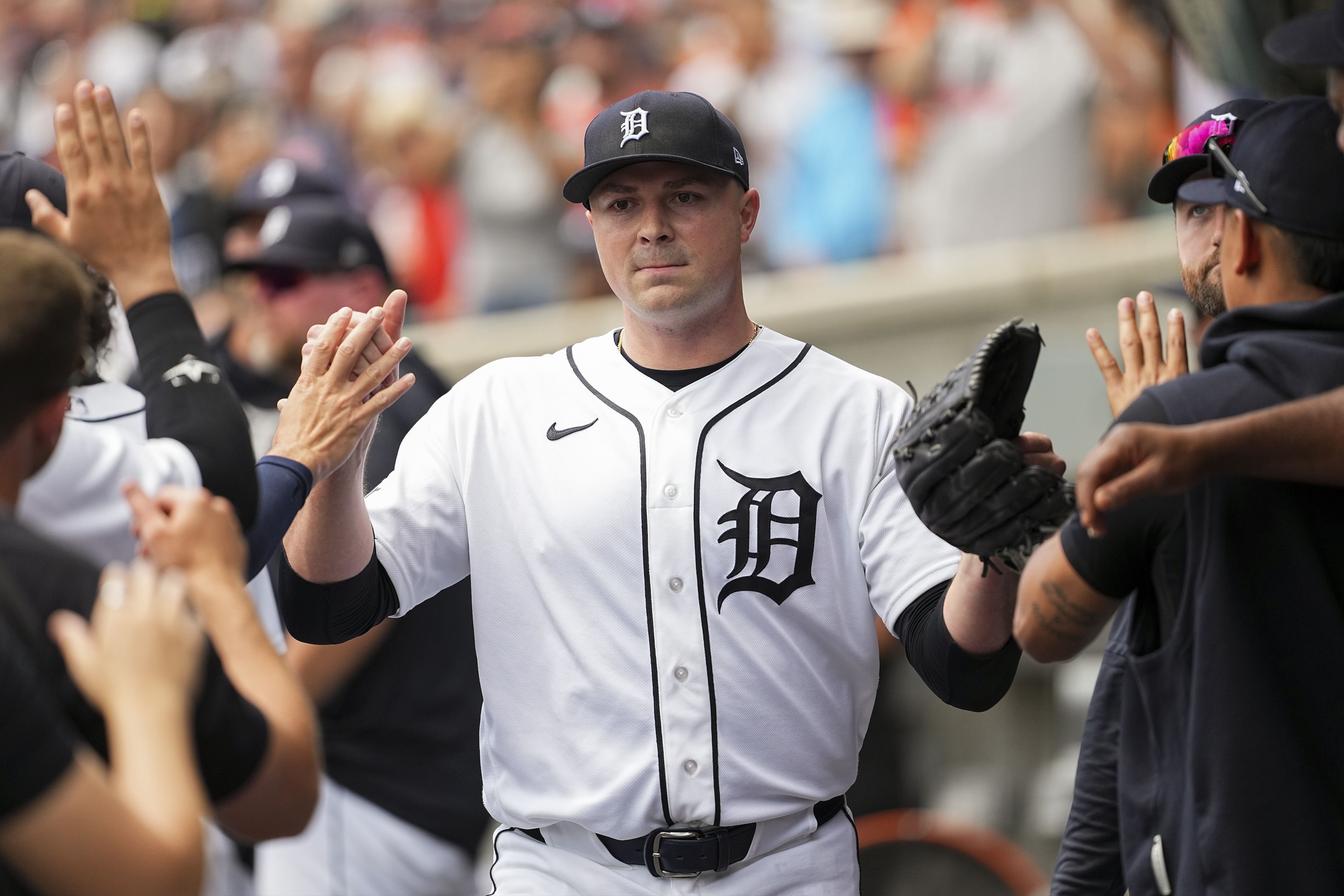 Baseball player in a Detroit Tigers uniform wears a black cap with a white D, white jersey with black piping, and high-fives teammates as a crowd watches in the blurred stands.