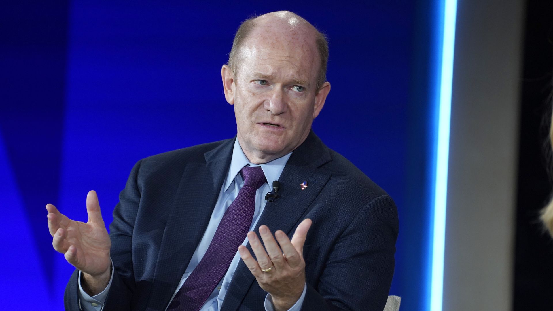 Sen. Chris Coons, wearing a blue suit and holding up his hands while sitting in a chair in front of a blue backdrop.