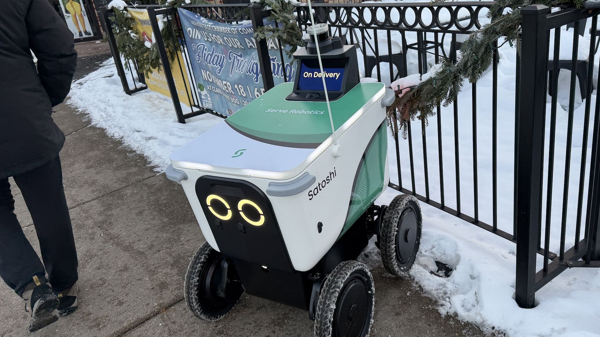 Green and white delivery robot named Satoshi on four wheels with glowing eyes, parked on snowy sidewalk near black metal fence decorated with garland, one person walking away.