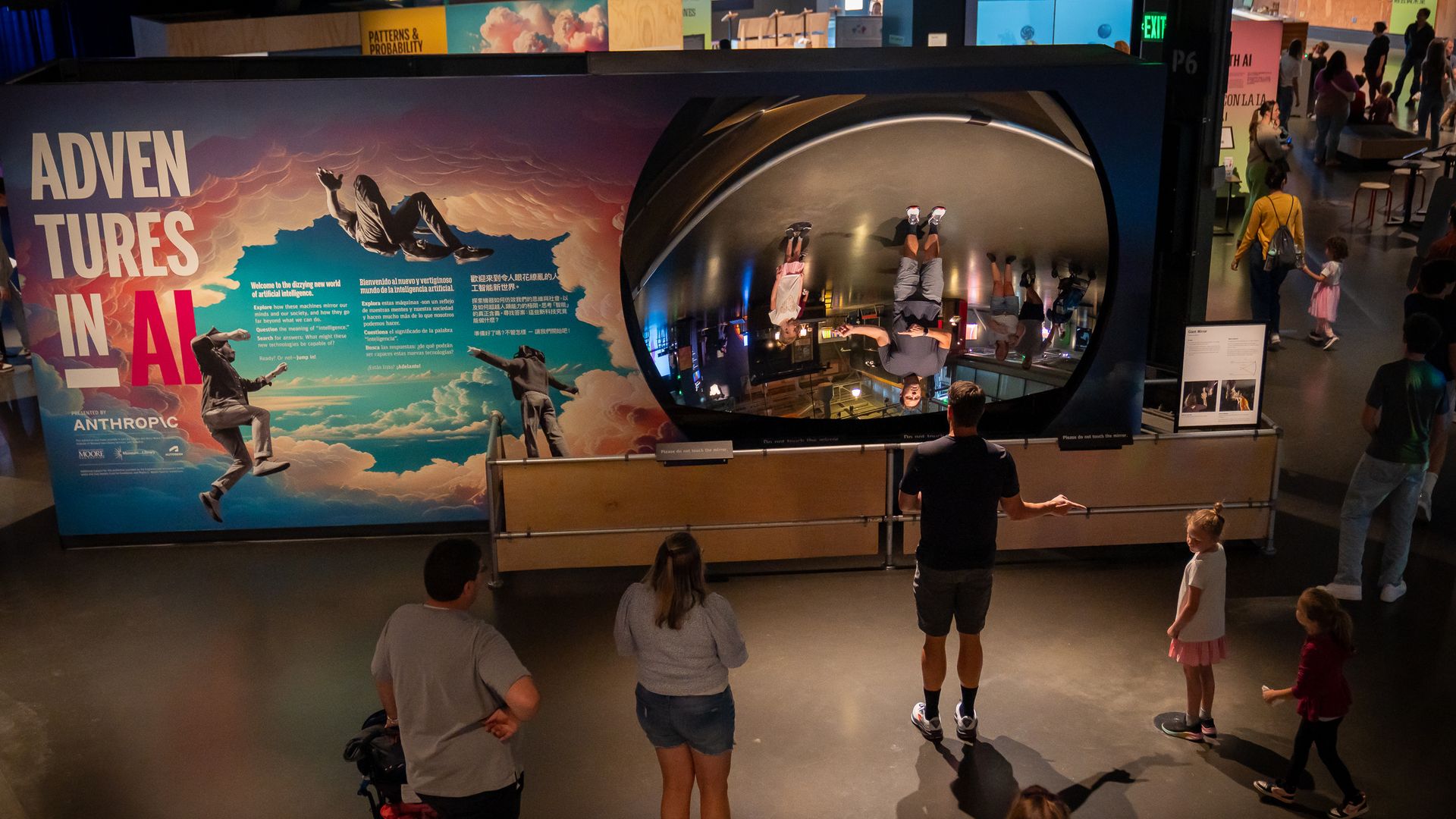 Museum exhibit titled "Adventures in AI" with colorful cloud artwork and text in multiple languages. People, including children, observe a large curved mirror reflecting visitors upside down.