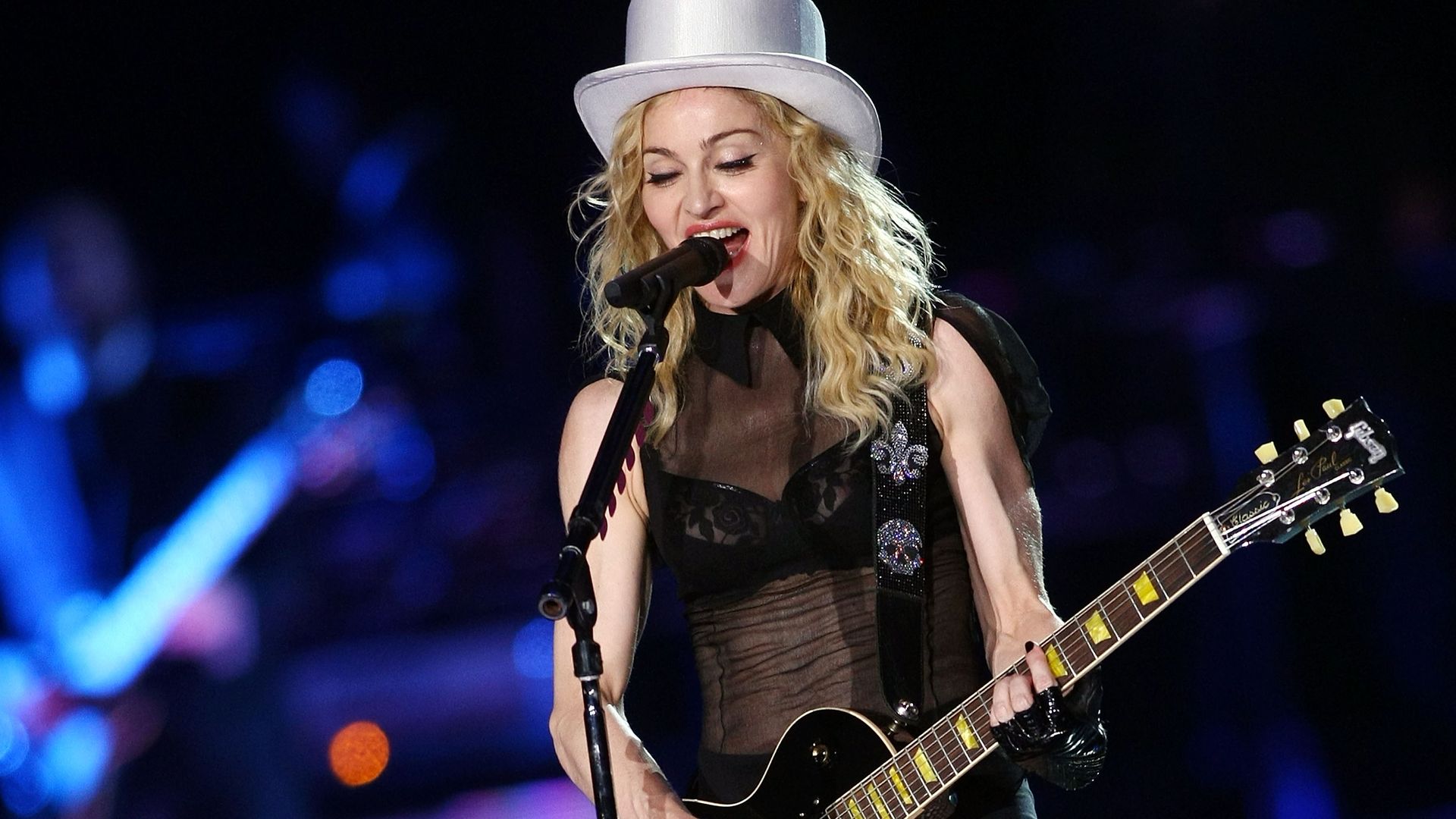 Pop icon Madonna sings into a microphone while playing guitar in a hat. 