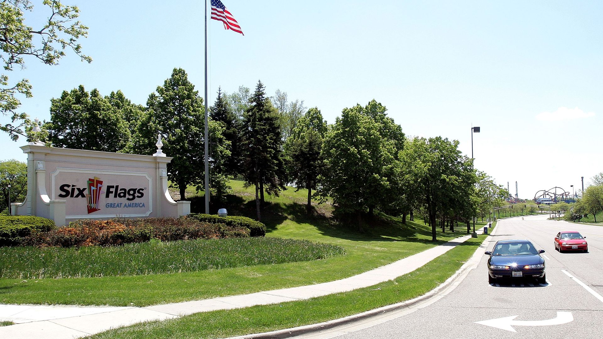 Six Flags Great America entrance sign with a tall flagpole and waving American flag, surrounded by green trees, a curved road, and a clear blue sky.