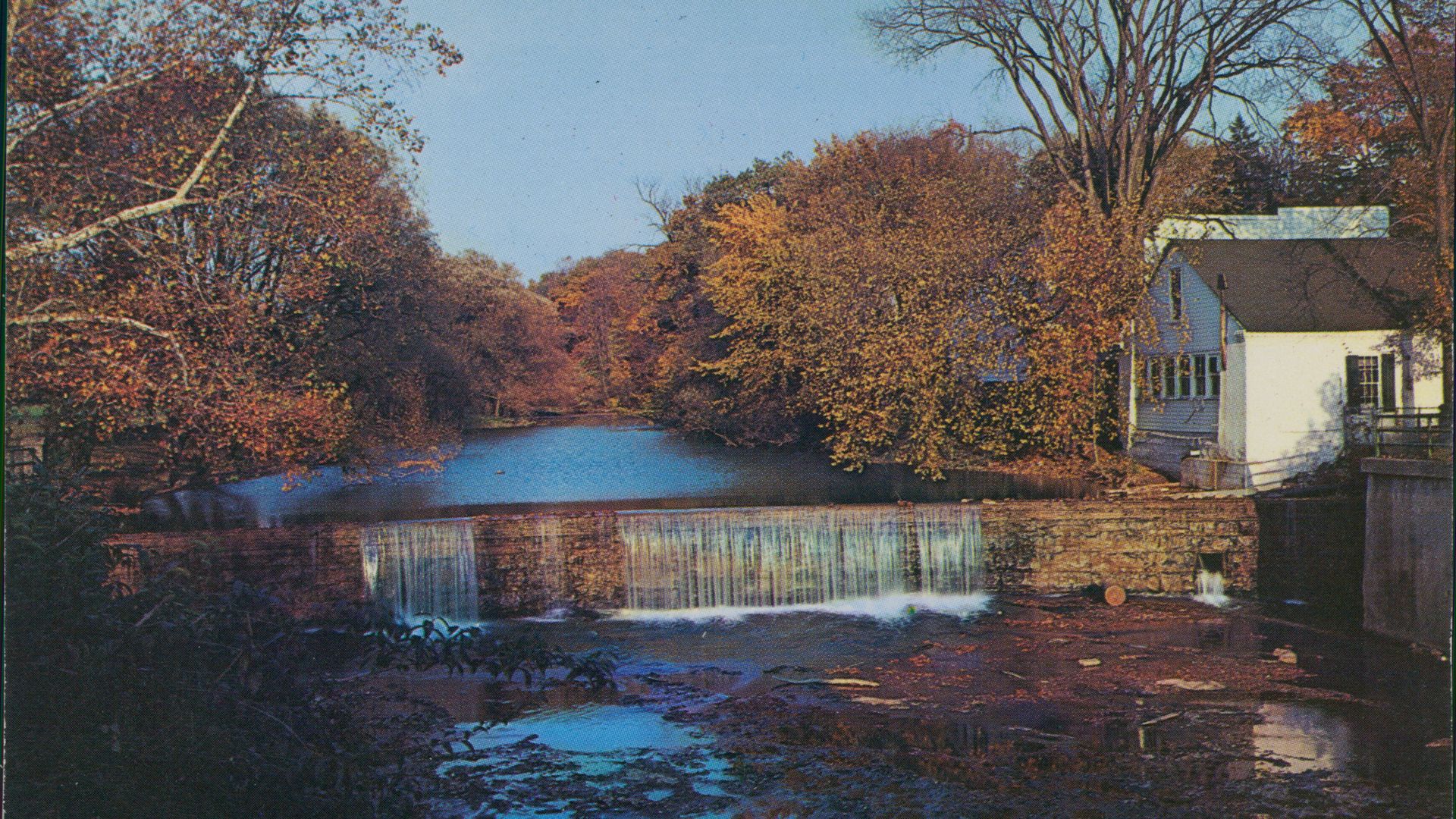 Postcard of quaint waterfalls with autumn trees on banks and white house on righthand side.