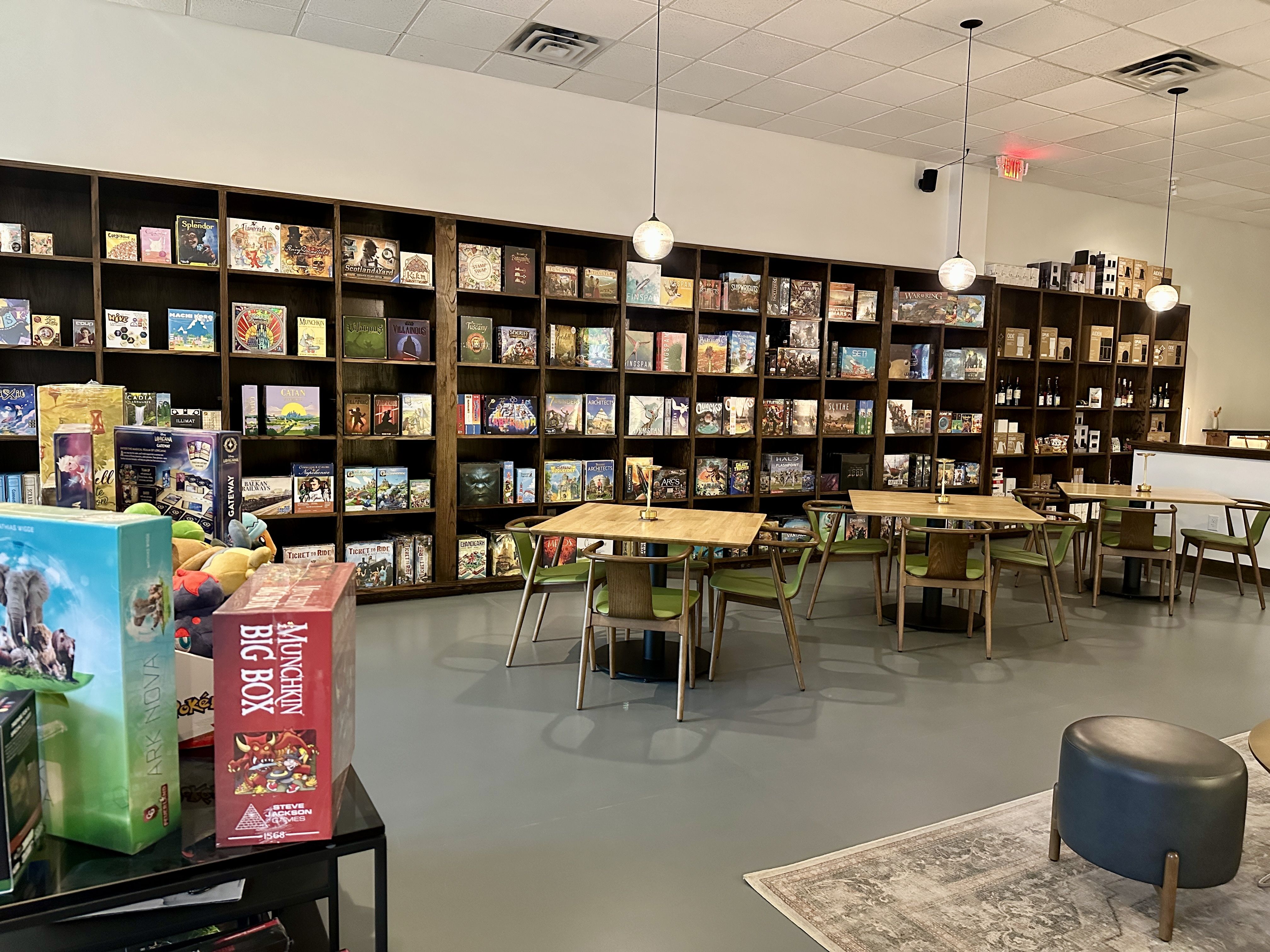 Interior of a board game cafe with wooden tables, green chairs, hanging pendant lights, shelves filled with numerous colorful board games, and a black ottoman on a patterned rug.