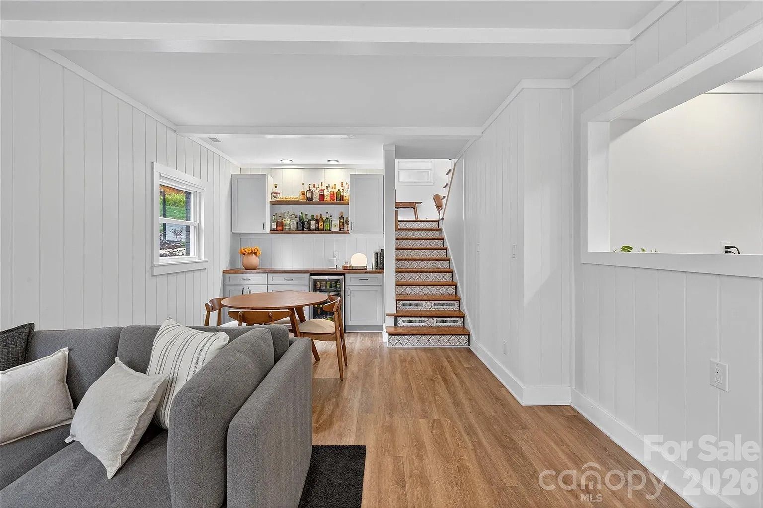 Cozy living space with gray sofa, wooden dining table, bar area with bottles, white paneled walls, patterned stair risers, and wood flooring under bright lighting.