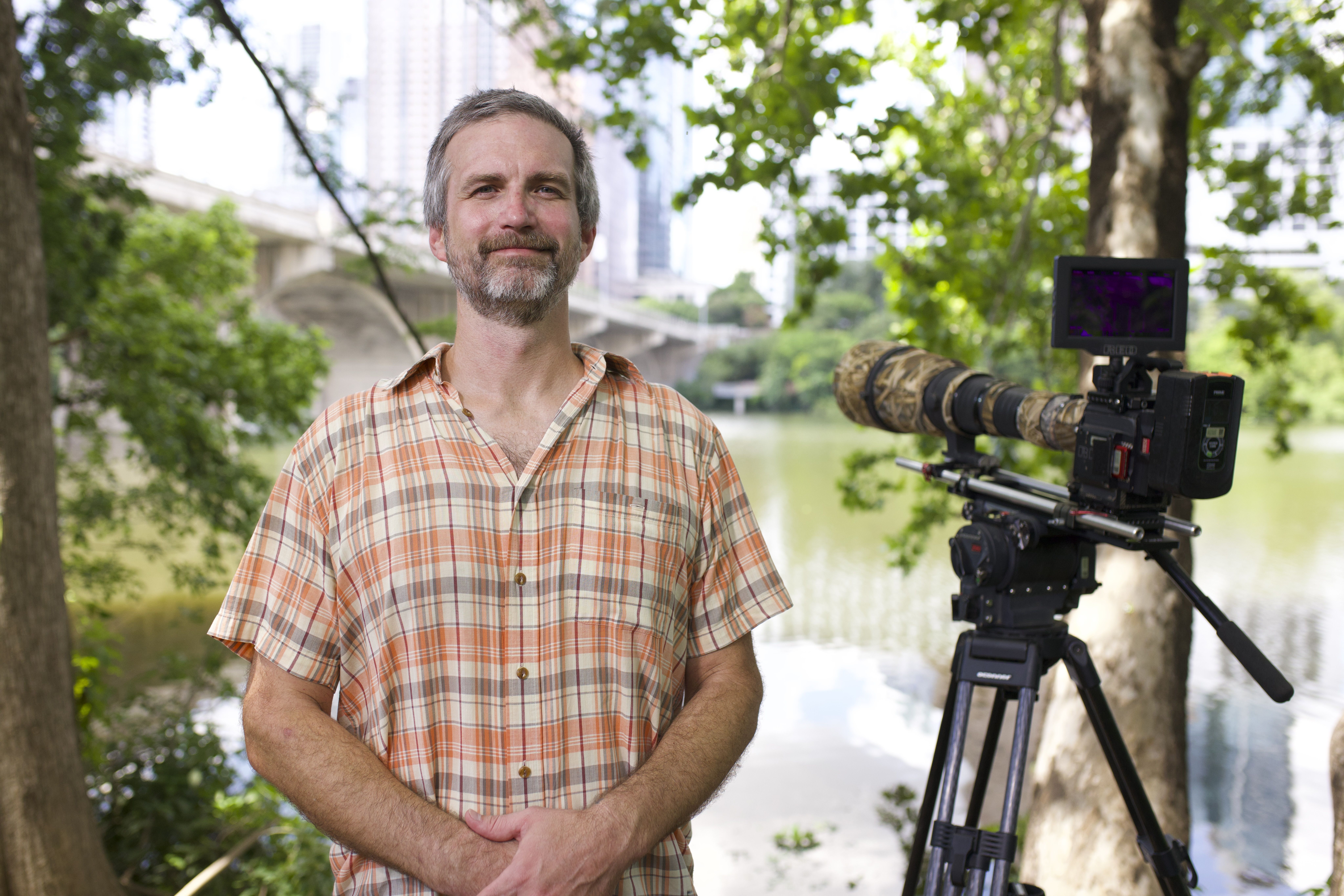 Smiling bearded man in an orange plaid shirt stands beside a large video camera on a tripod outdoors by a river, with trees and city buildings in the background.