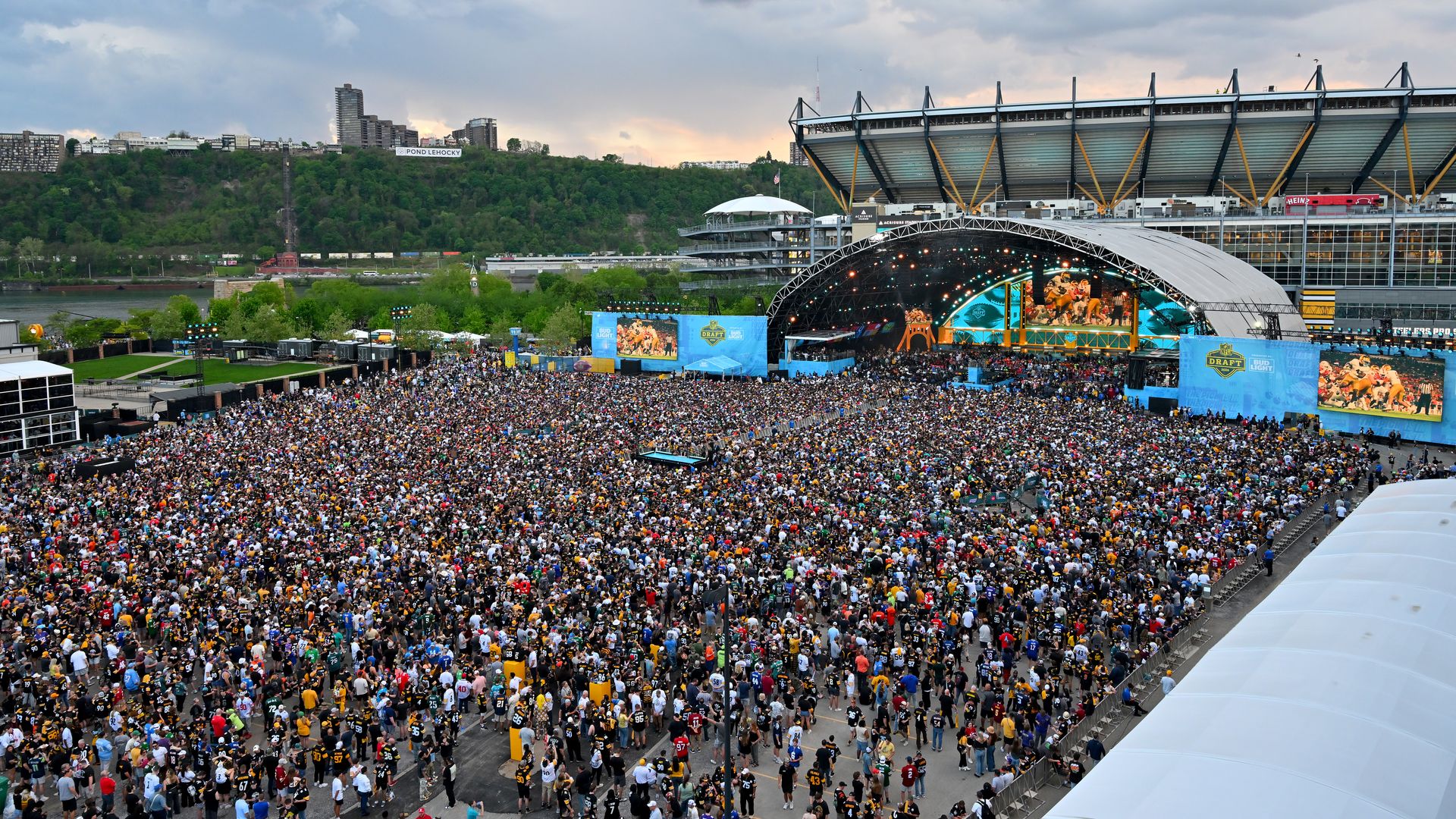 Massive densely packed crowd, a brightly lit stage with screens, blue banners, and a river and green hillside beyond, under a cloudy sky.