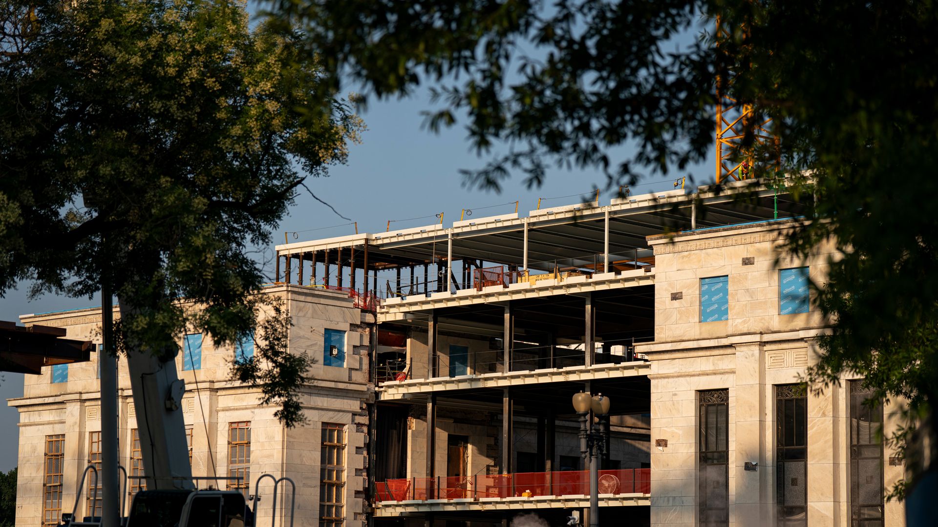 Partially constructed Federal Reserve building with a marble facade and steel framework, surrounded by construction fencing and equipment, framed by tree branches under a clear sky.