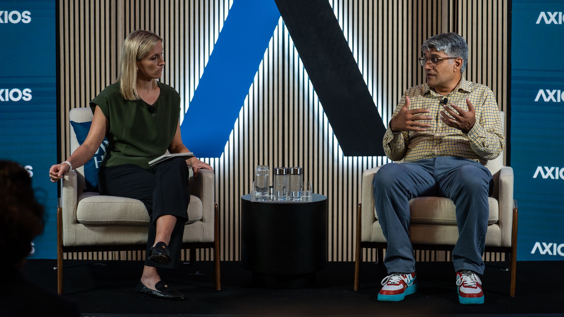 Two speakers sit in chairs: a woman in green on the left and a man in plaid on the right, gesturing as they talk. A blue-black geometric backdrop with AXIOS branding and a glass table between.