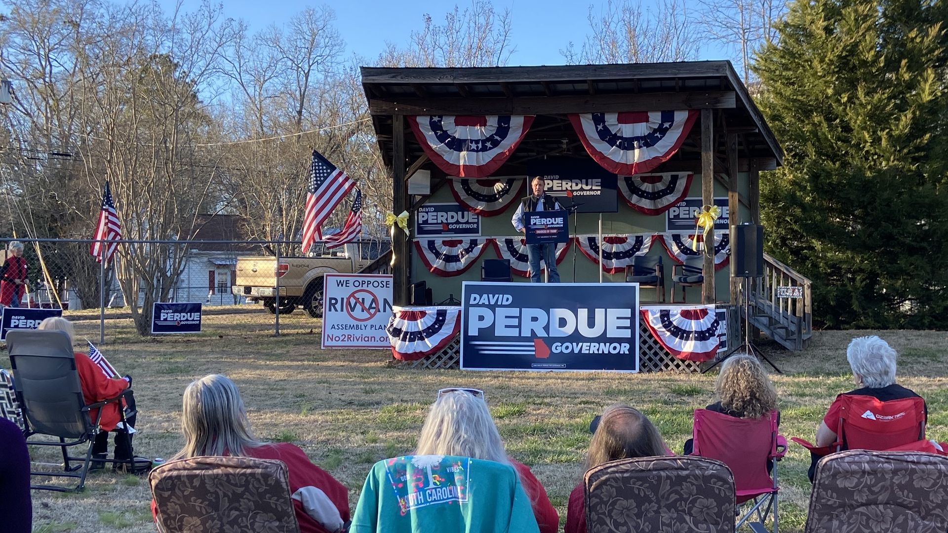 Perdue speaking on a stage to people in lawn chairs