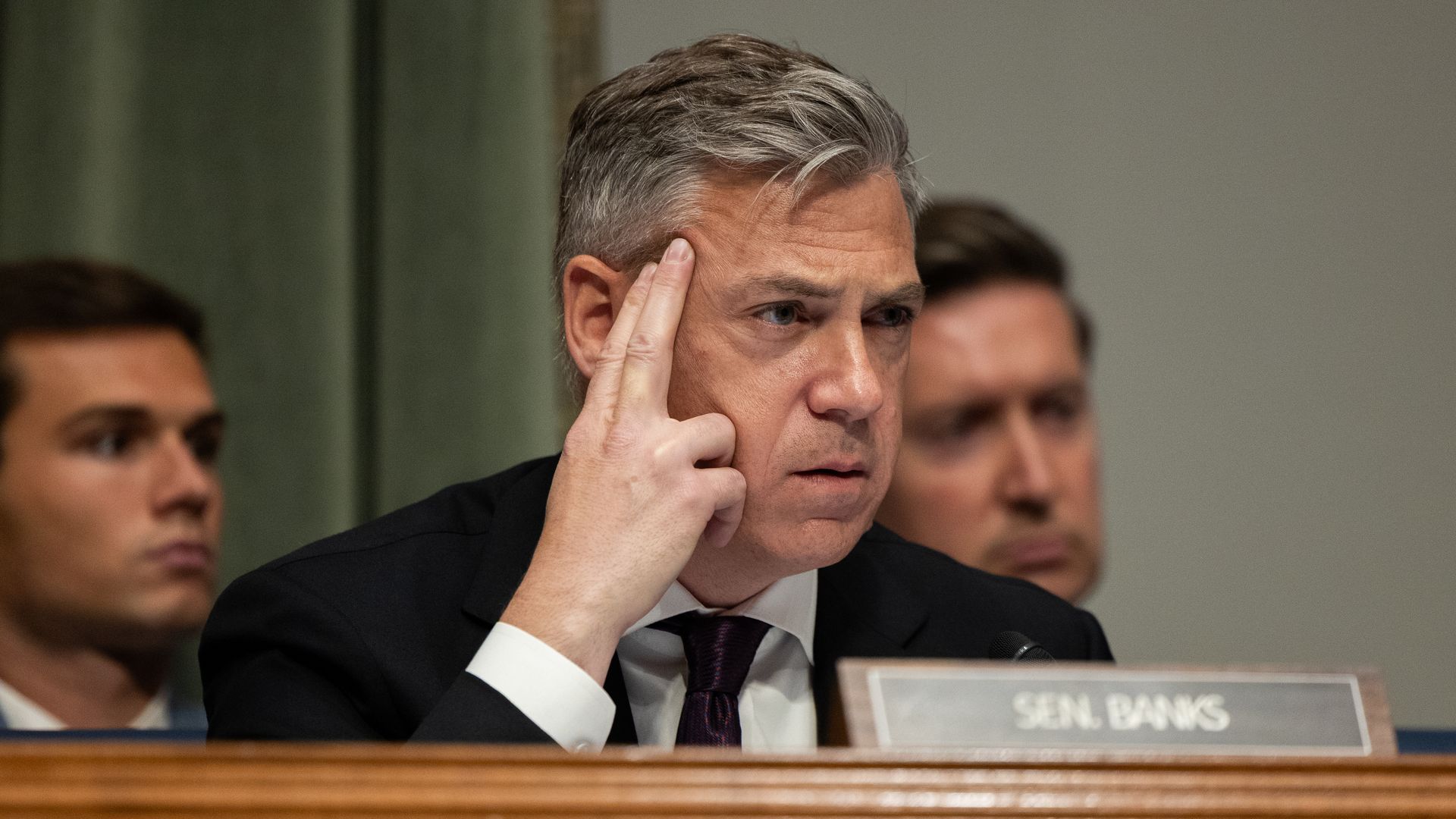 A man in a suit and tie identified as Sen. Banks touches his temple with two fingers, looking pensive during a formal meeting with two blurred men in background.