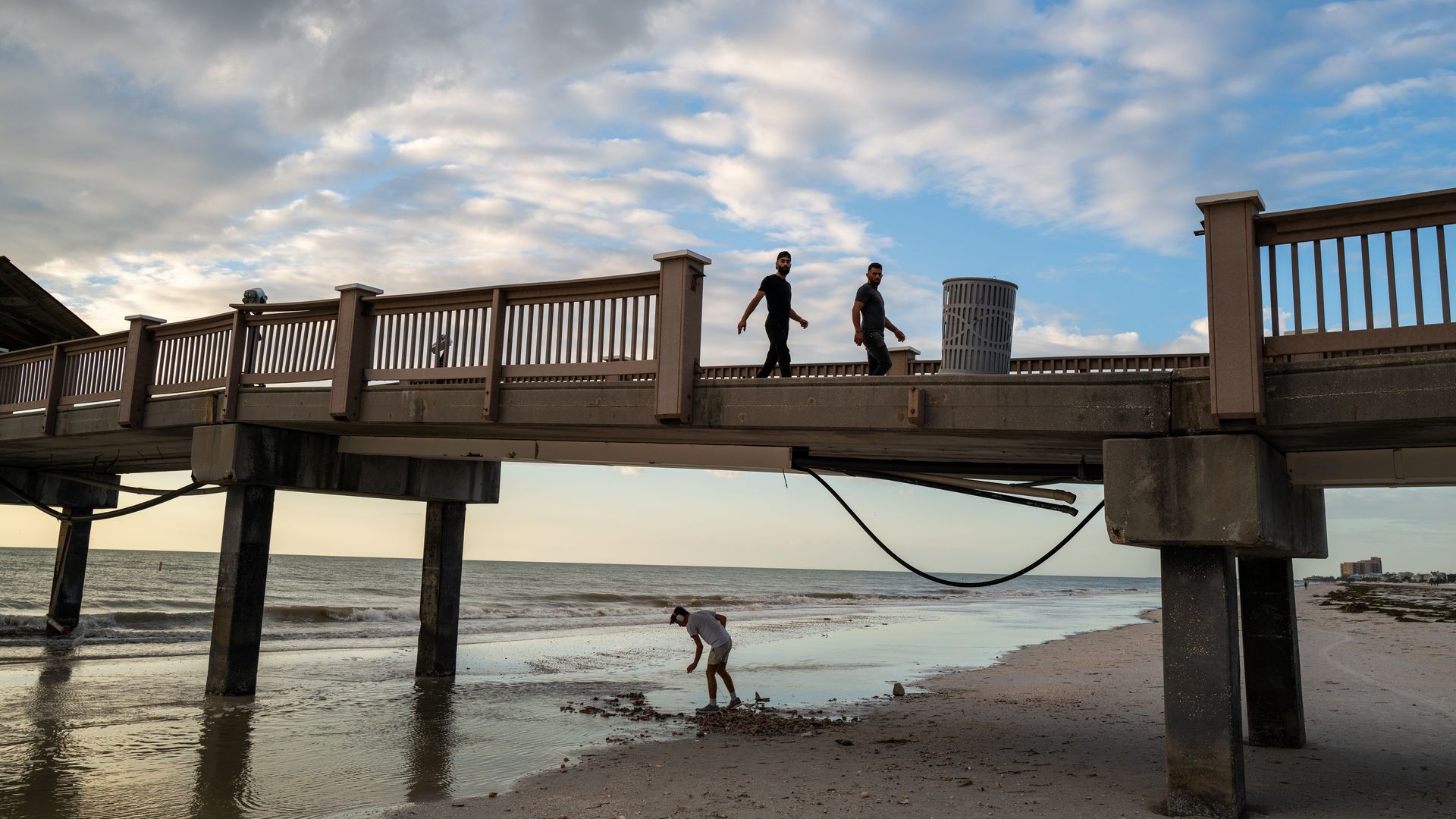 Two people walk on a beach boardwalk. A third looks at sand beneath the boardwalk.