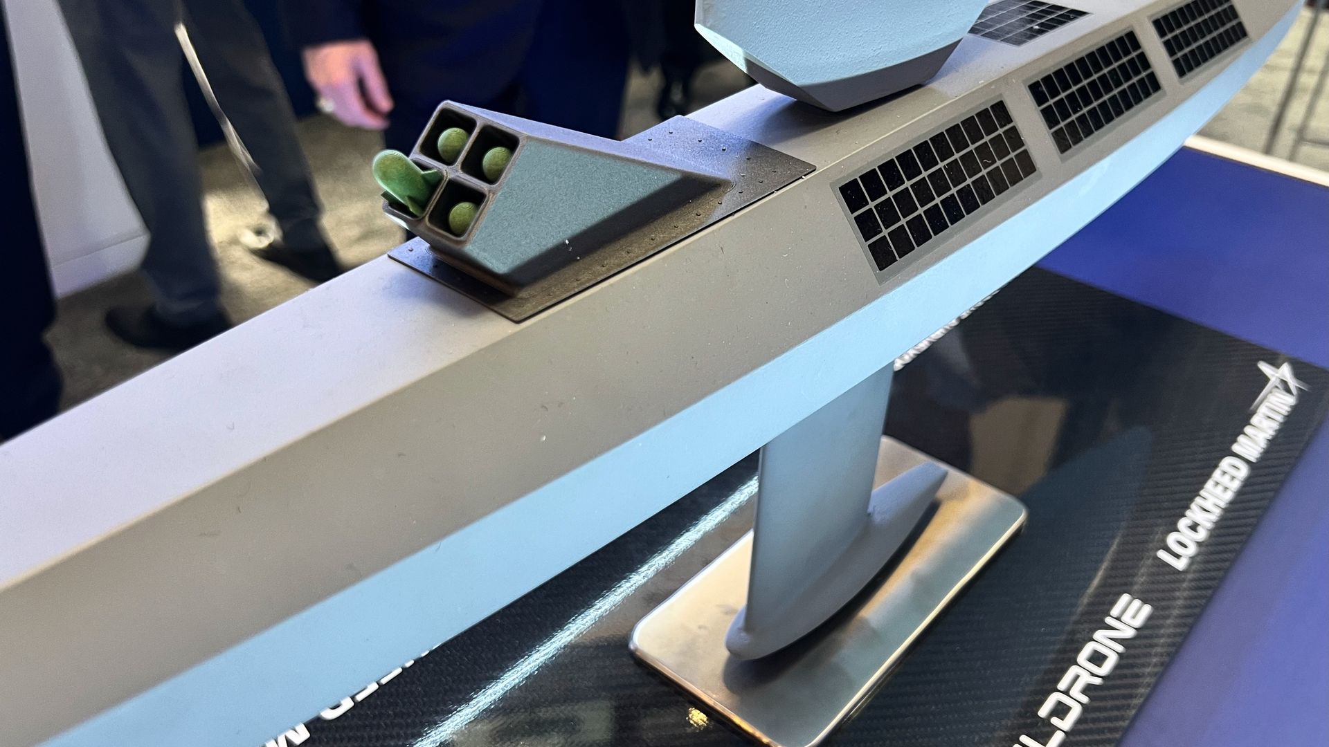 Close-up of a modern grey boat model with black solar panels on the deck and green cylindrical components at the rear, displayed on a black base labeled "Saildrone Lockheed Martin."