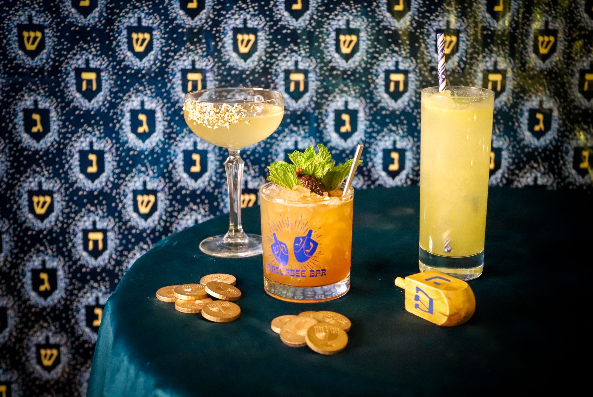 Three cocktails, near a dreidel in front of blue wallpaper with dreidels with Hebrew letters as part of Macabee, the Hannukah pop-up bar in Cambridge.