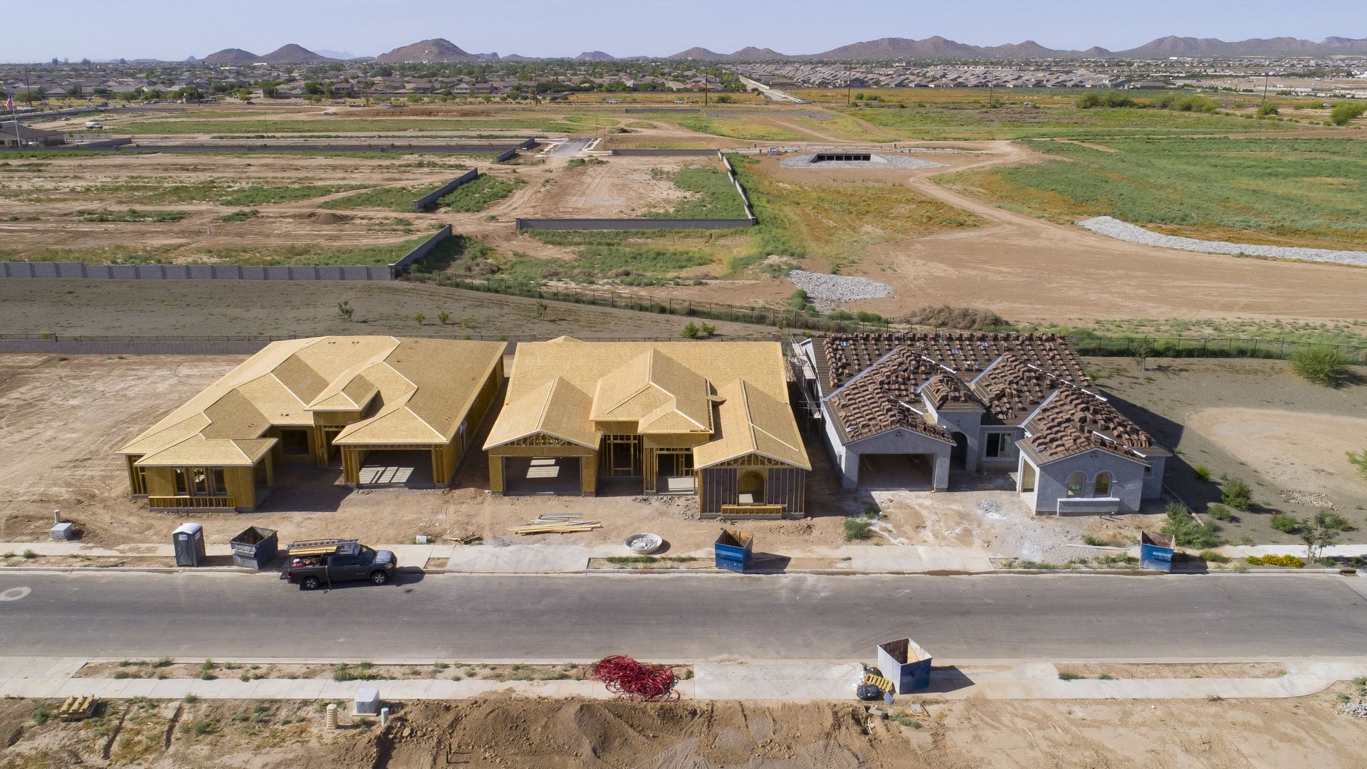 Aerial view of three houses under construction along a street: left and middle with wooden framing, right nearly finished with gray exterior and red-tiled roof; desert landscape and distant hills.
