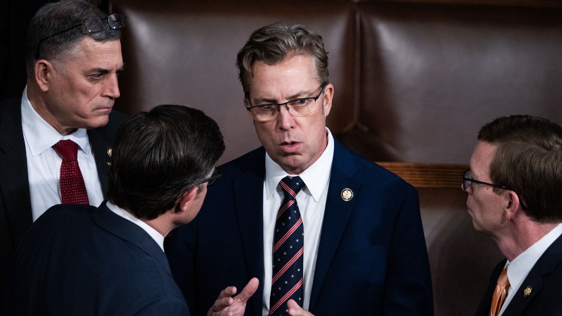 Rep. Andy Ogles, wearing a blue suit and speaking to three colleagues while standing in front of two rows of brown leather chairs.