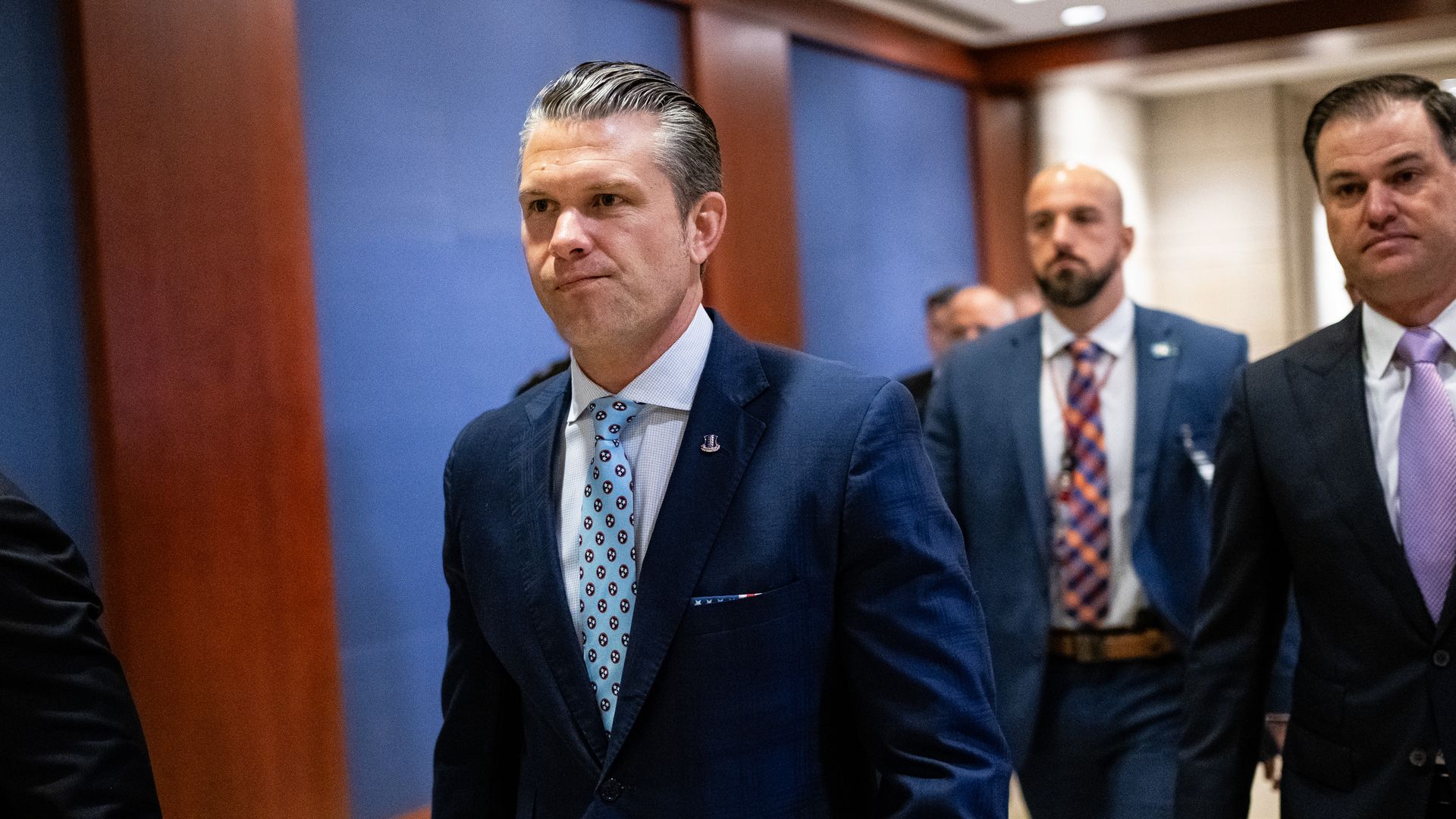 Secretary of Defense Pete Hegseth, wearing a blue suit and walking through a blue and wood-paneled hallway while flanked by aides and security.