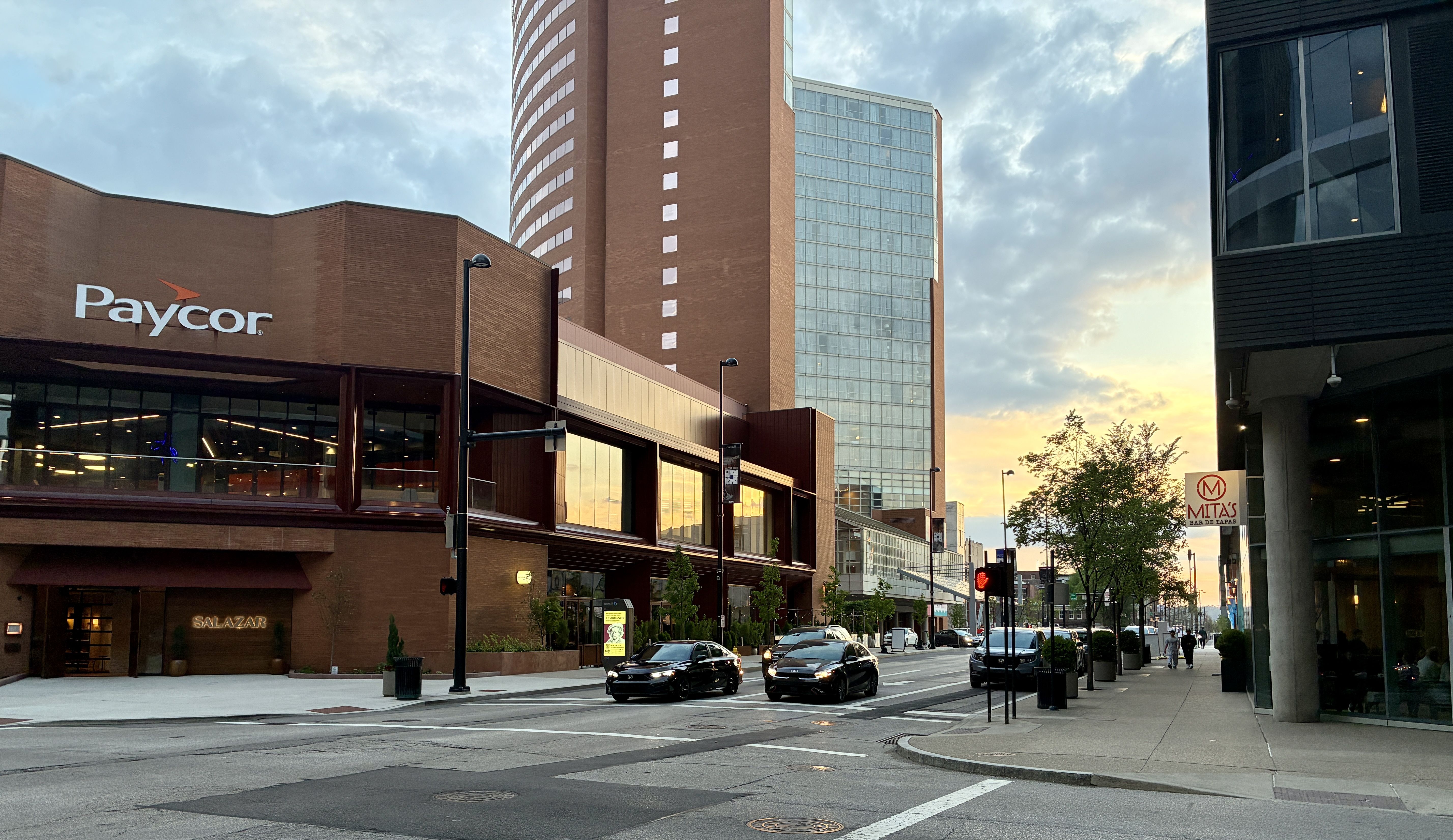 City street scene at sunset: a brick Paycor building on the left, a tall brick-and-glass tower in the center, and a modern dark building on the right. Cars and pedestrians along the curb.