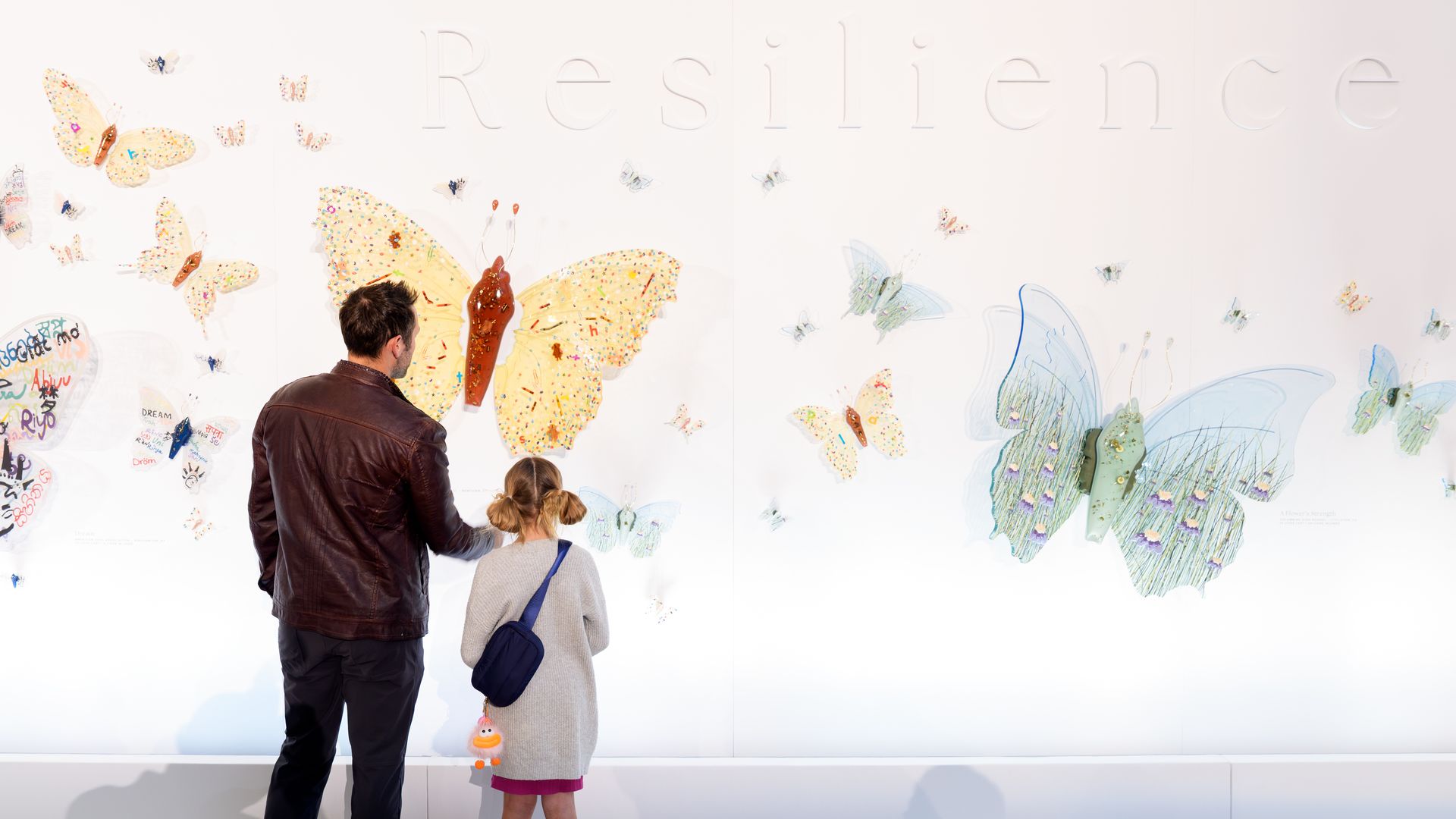 A man in a brown jacket and a girl in a gray sweater stand before a white wall art exhibit featuring large colorful butterflies and the word "Resilience" embossed on the wall.