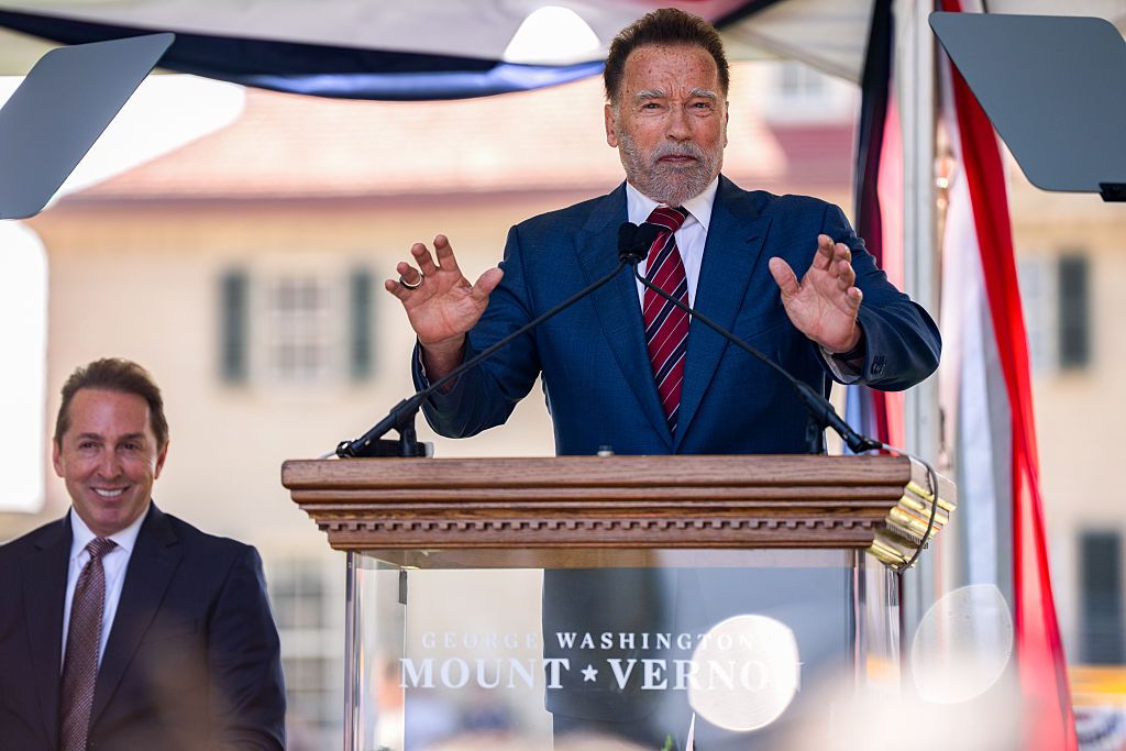 Arnold Schwarzenegger, wearing a blue suit and red, white and blue tie, stands at a podium with two microphones. His hands are out in front of him. 