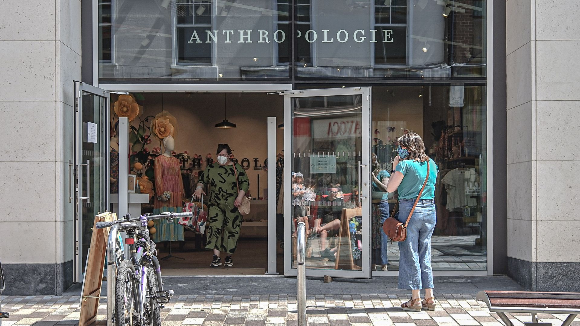 Anthropologie store entrance with glass doors open, a woman in green dress and mask inside, and another woman in blue top and mask standing outside. Bicycles parked nearby on tiled pavement.