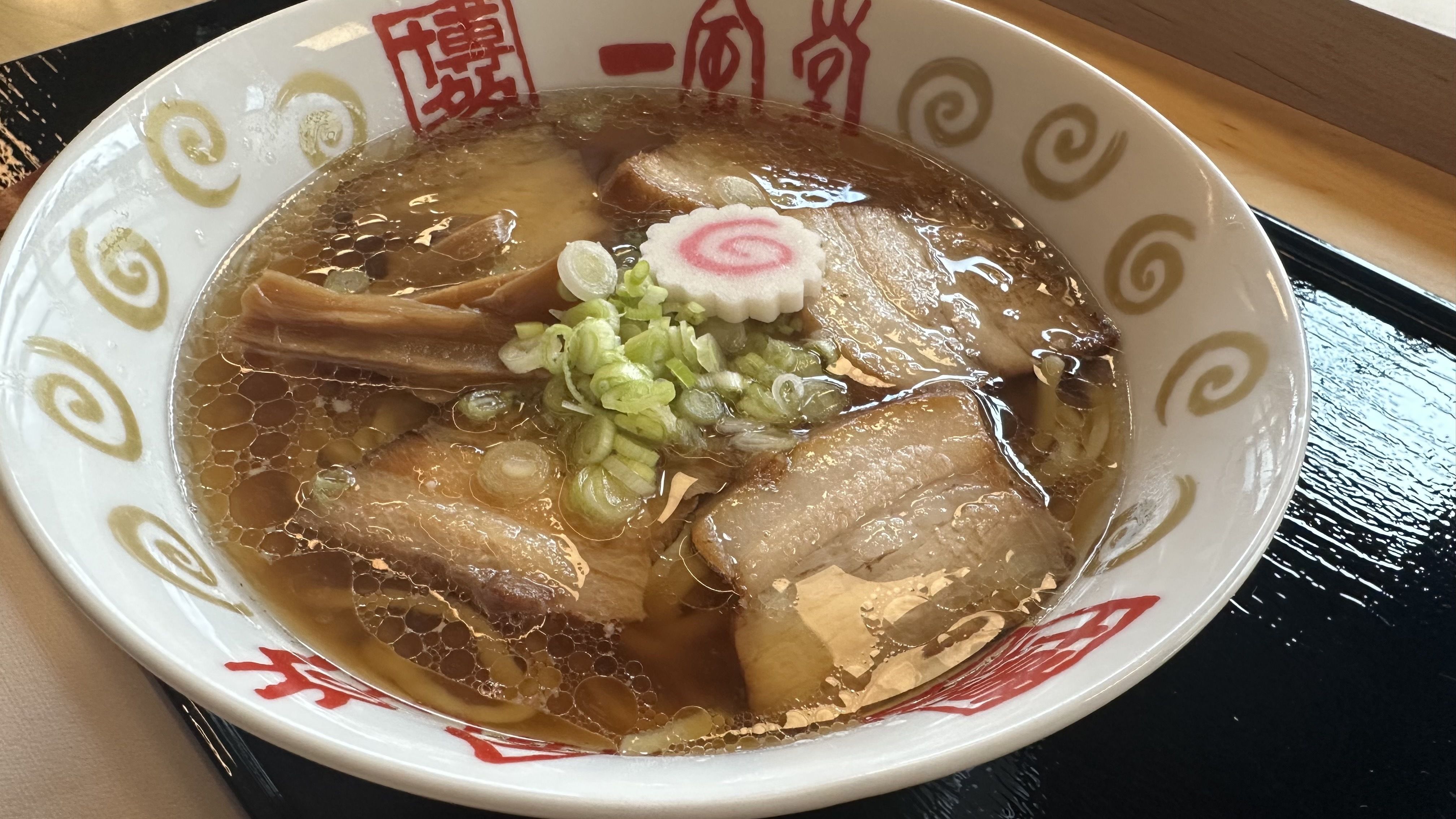Top-down view of a ramen bowl with rich brown broth, oil beads, sliced pork, bamboo shoots, chopped green onions, and a white narutomaki with a pink swirl. Red kanji and gold swirls decorate the rim.