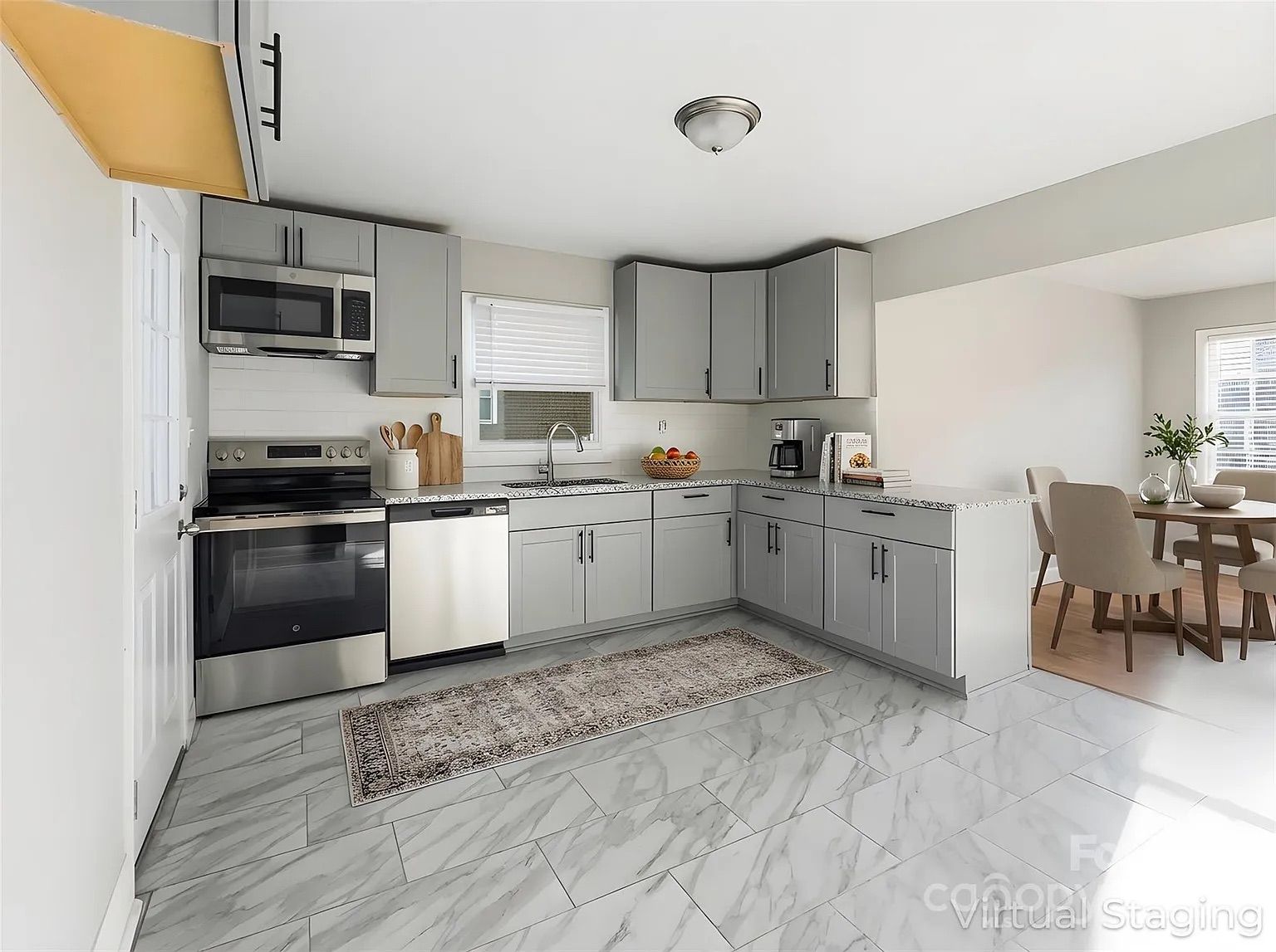 Modern kitchen with gray cabinets, stainless steel appliances, white walls, marble tile floor, a rug in front of sink, and adjacent dining area with beige chairs and wooden table.