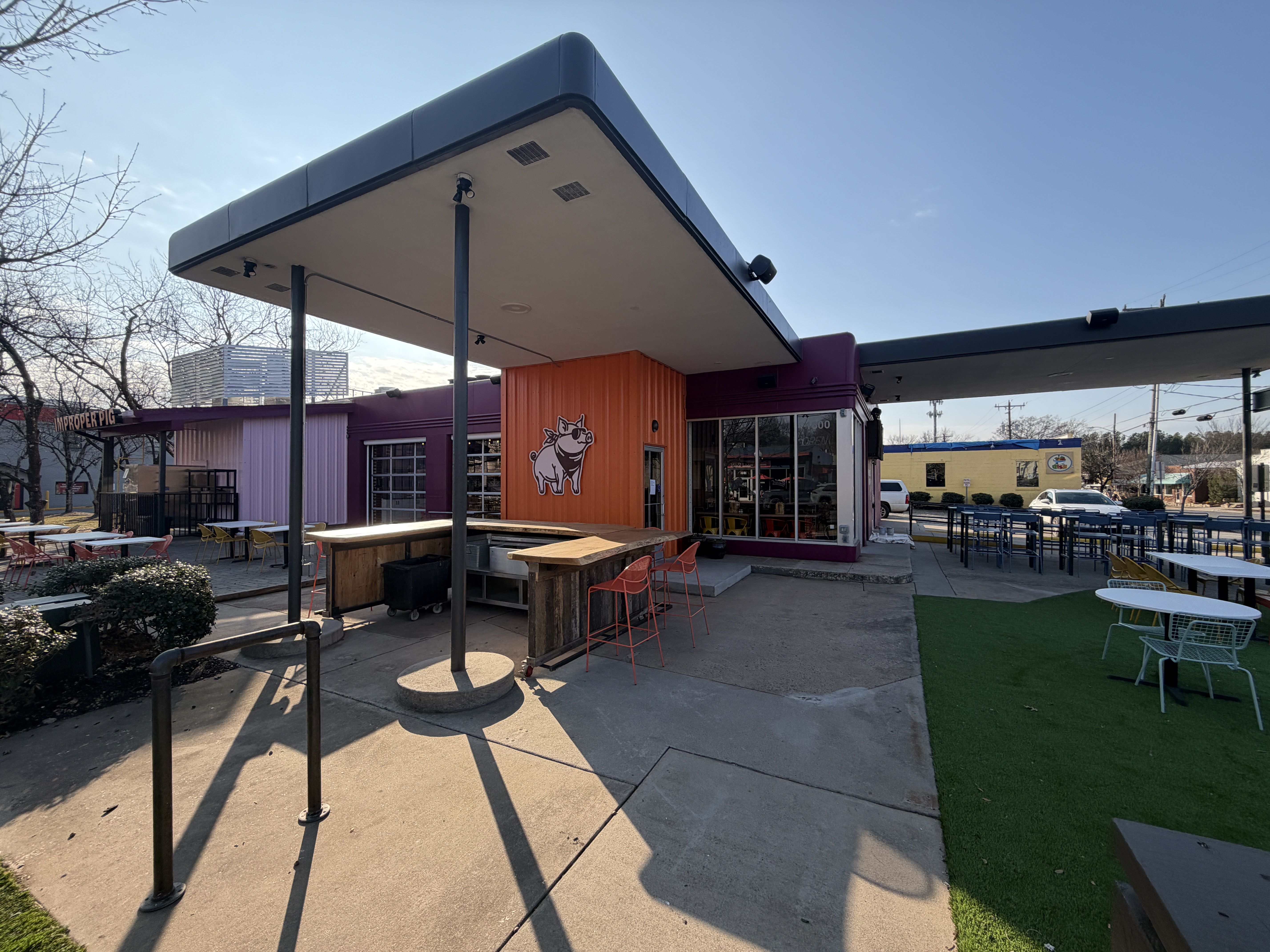 Outdoor seating area of a restaurant with colorful chairs and tables, a purple and orange building featuring a cartoon pig logo, under a clear blue sky.