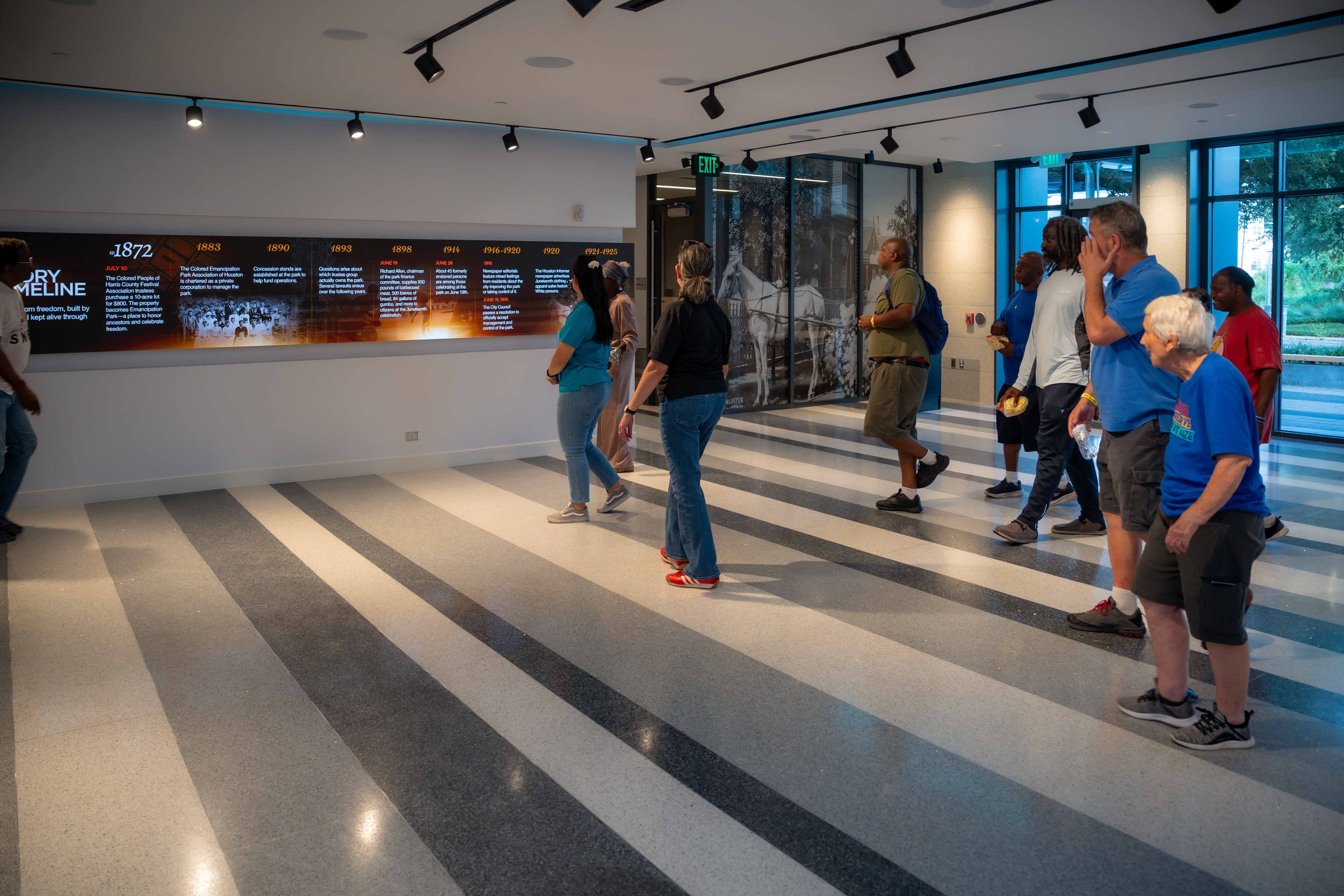People in a modern museum hallway study a long, illuminated timeline display along the left wall; track lighting overhead, glass doors and large windows on the right.