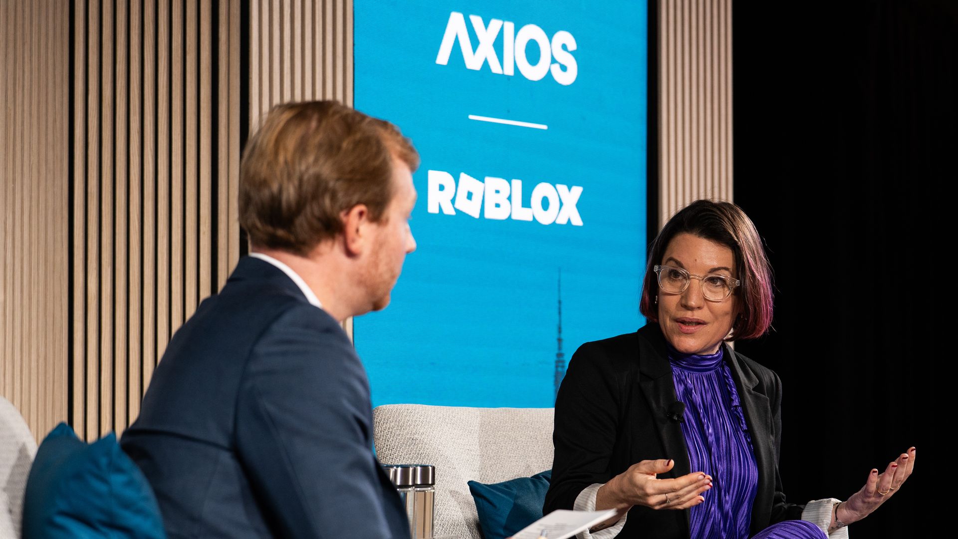 Two people in a discussion on a stage with a blue screen behind them displaying the logos "AXIOS" and "ROBLOX." The woman wears a purple dress and black jacket; the man faces her holding notes.