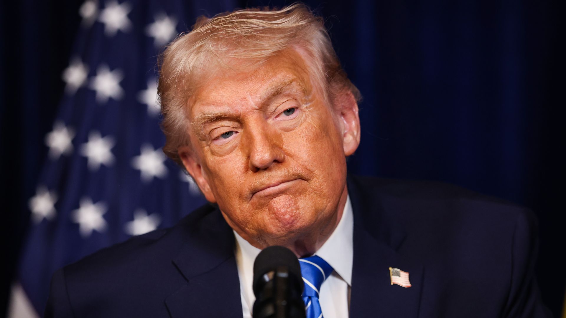 President Trump — wearing a navy suit, a blue tie, a white collared shirt and an American flag pin on his lapel — frowns as he appears in front of a microphone with an American flag behind him.