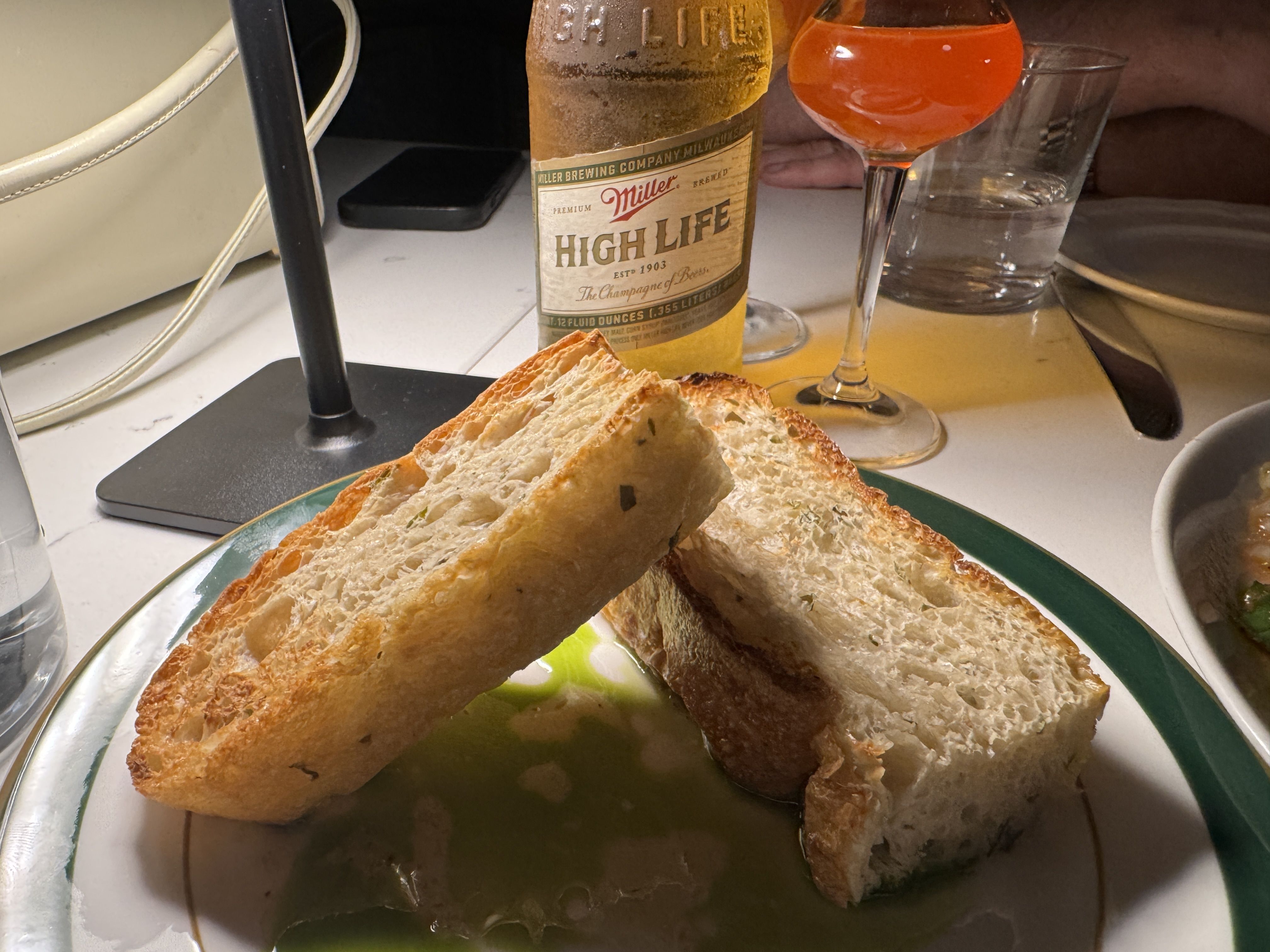 Two toasted bread slices on a plate with green oil or sauce, a Miller High Life beer bottle, a glass of orange drink, and a glass of water on a lit table indoors.