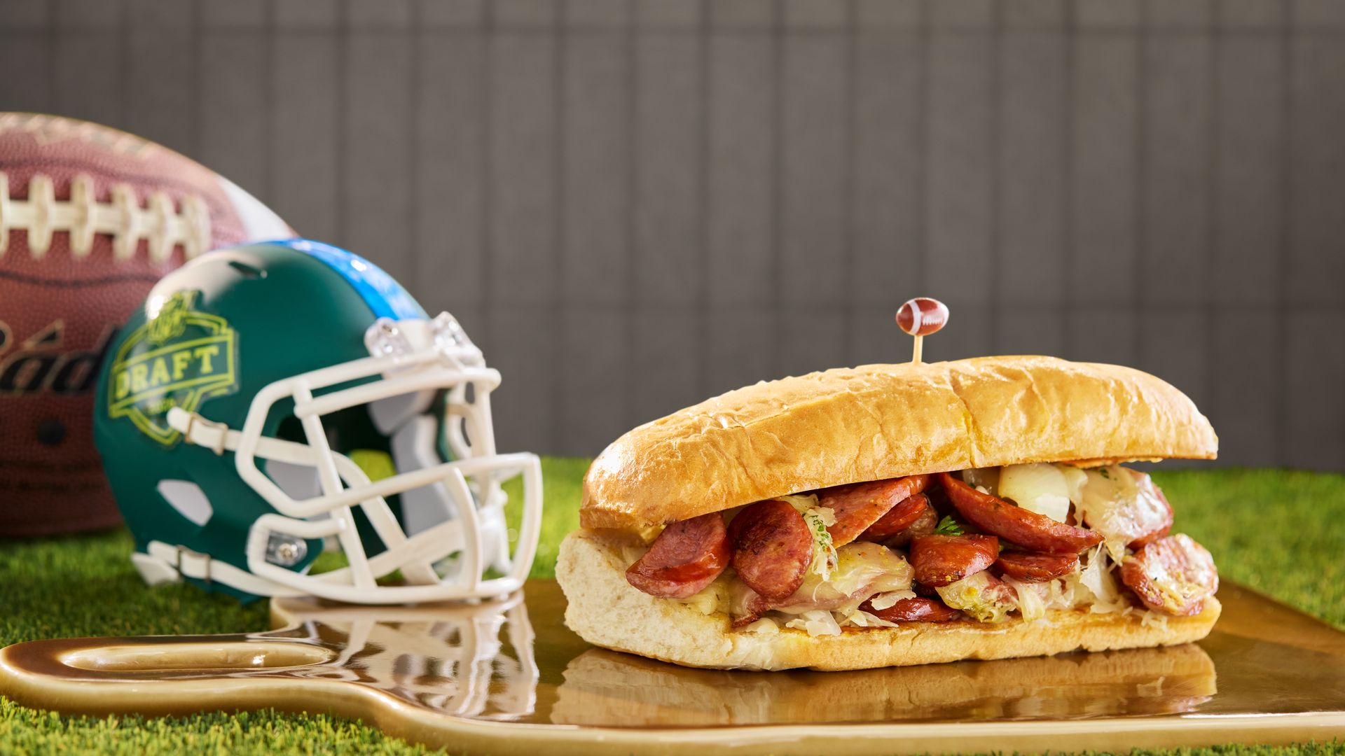 A close-up shows a sausage-filled sandwich on a gold plate, with a tiny football on a toothpick. A green football helmet with a DRAFT badge rests on artificial turf near a gray fence.