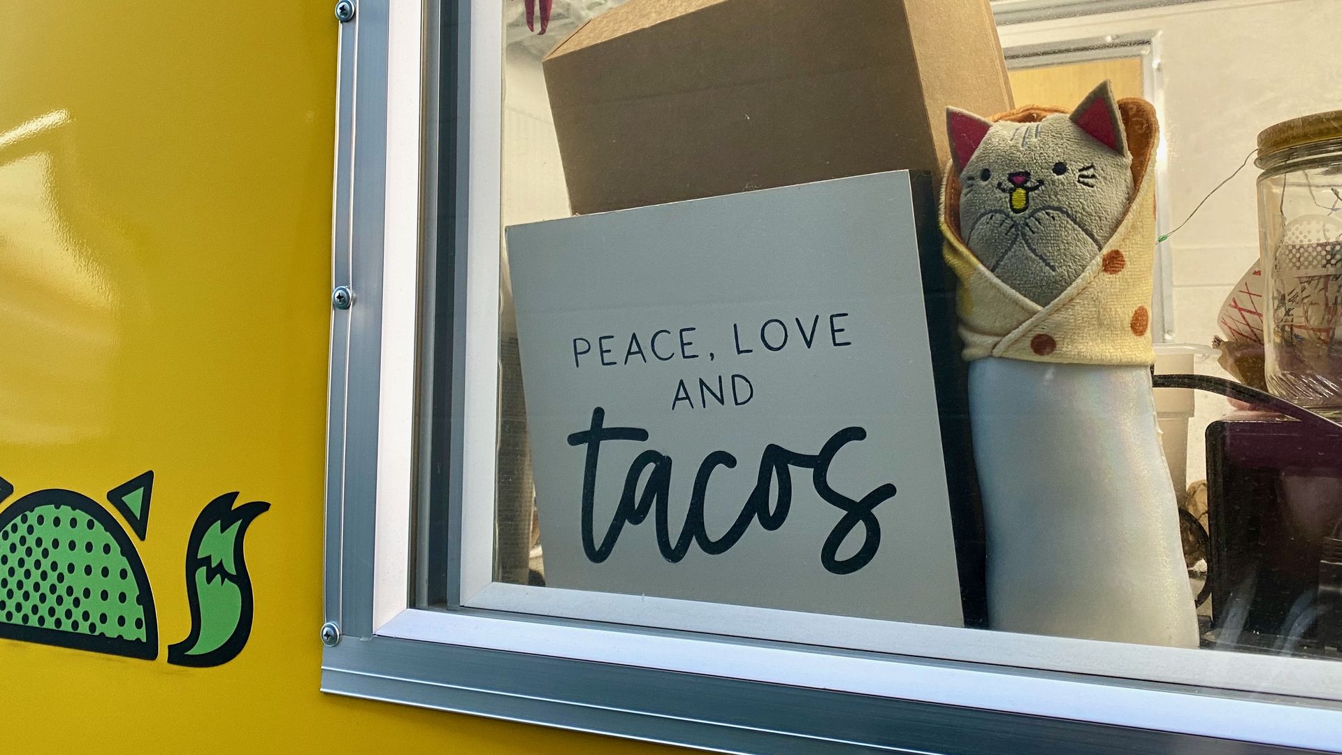 A sign at a taco stand counter reading "Peace, Love and Tacos" with a small cat toy. 