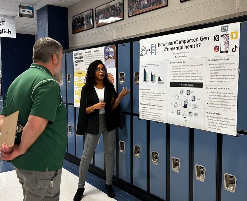 A student, a young woman, gestures toward a research presentation titled "How has AI impacted Gen Z Health?" as a teacher looks on. 
